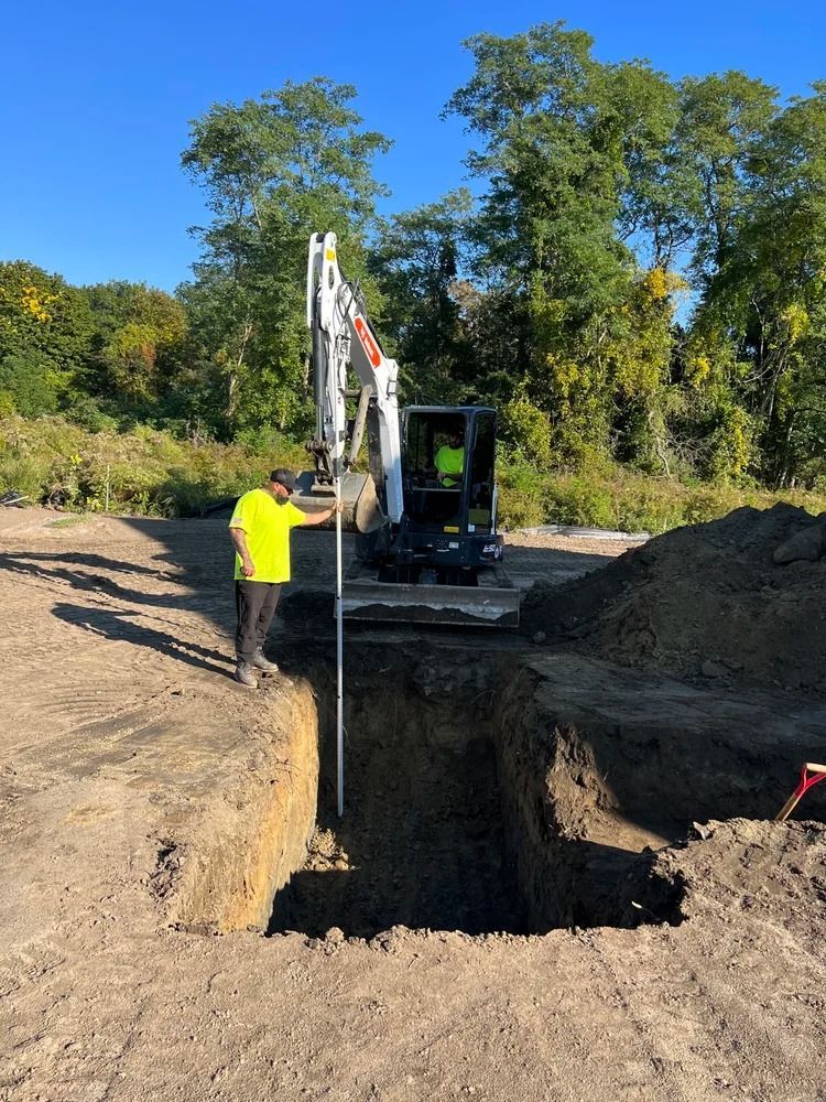 Man in yellow shirt measures depth of a rectangular excavation pit with a Bobcat excavator in background.