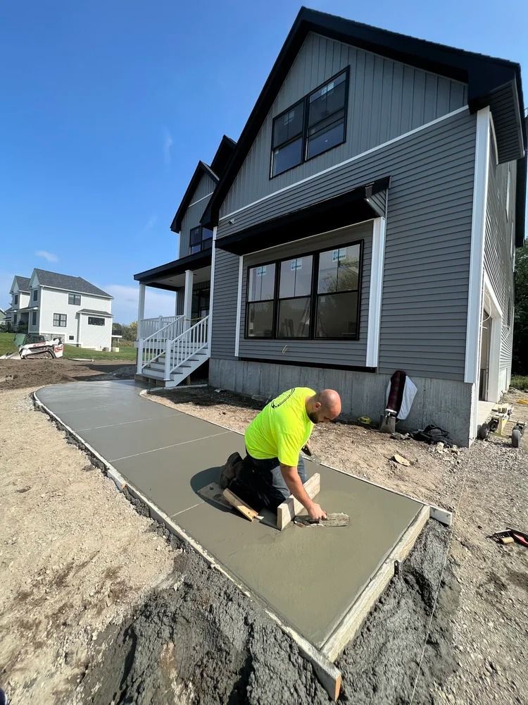 Man kneeling, smoothing wet concrete sidewalk in front of new gray house, sunny day.