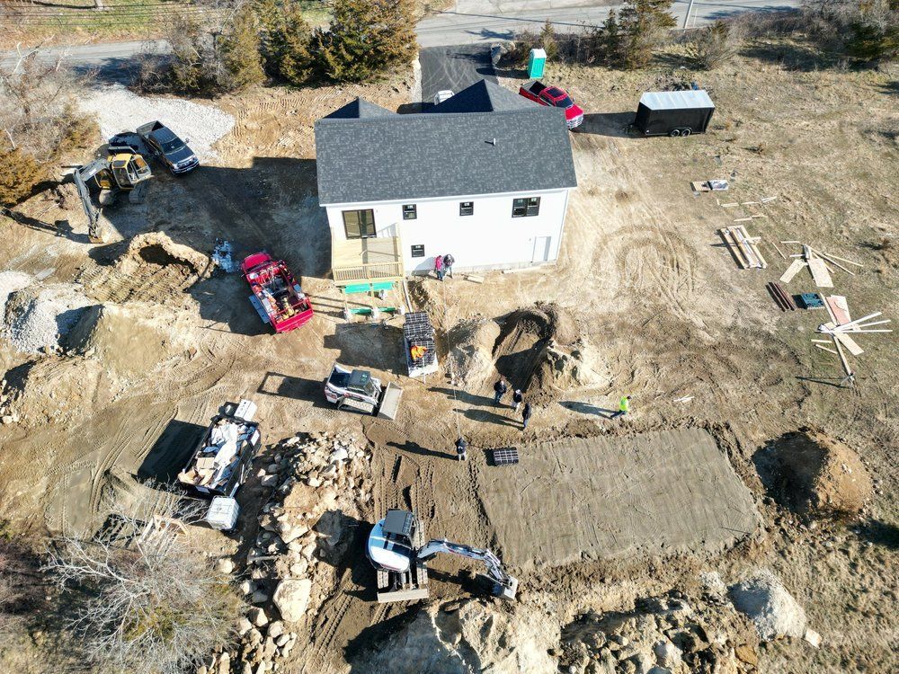 Aerial view of a building site with a partially constructed white house, construction equipment, and exposed earth.