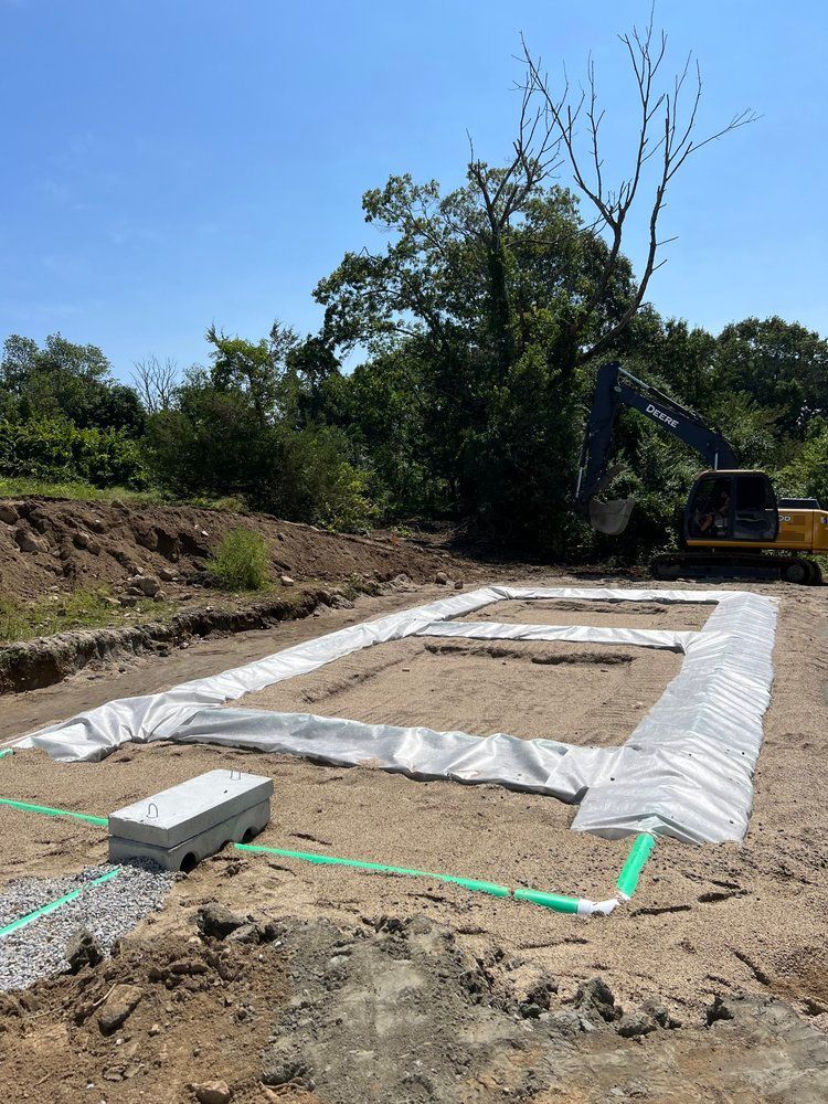 Construction site with a rectangular outline marked by white fabric and green piping. An excavator is in the background.