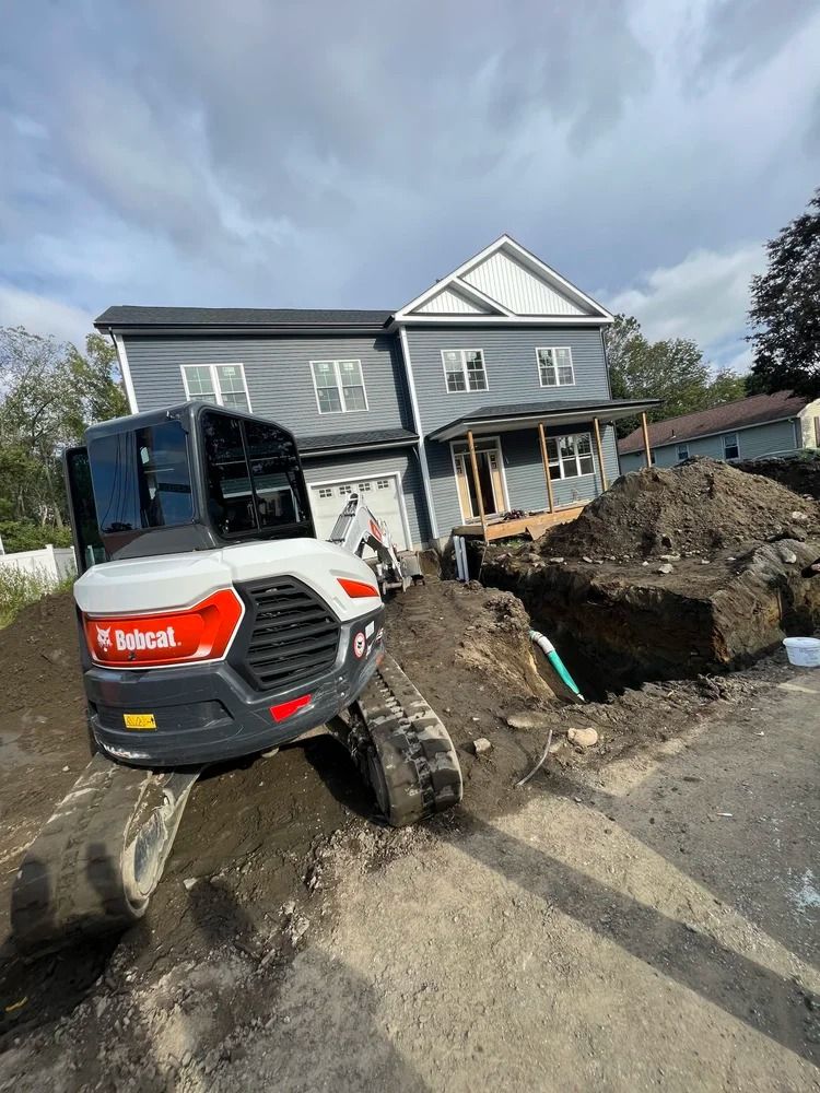 A Bobcat excavator digs near a two-story gray house under a cloudy sky. A construction site is visible.