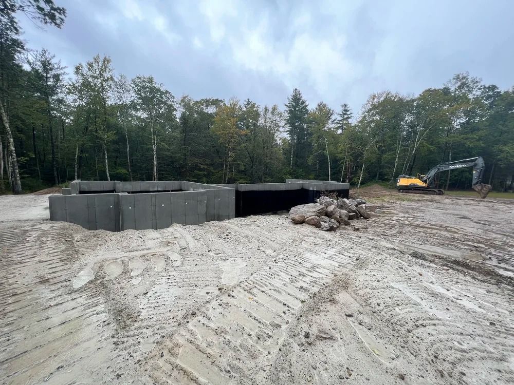 Construction site: concrete blocks in sand, excavator, trees in the background under cloudy sky.