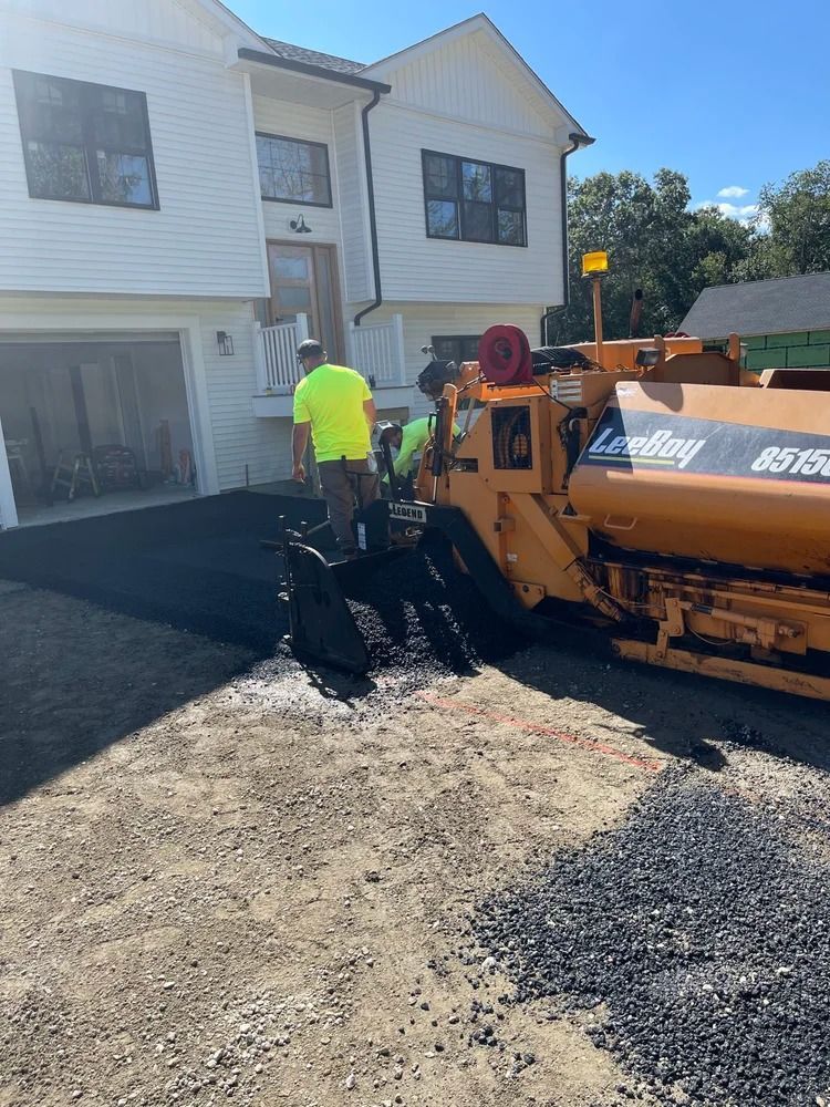 Workers paving a driveway in front of a white house with a paving machine, laying down black asphalt.