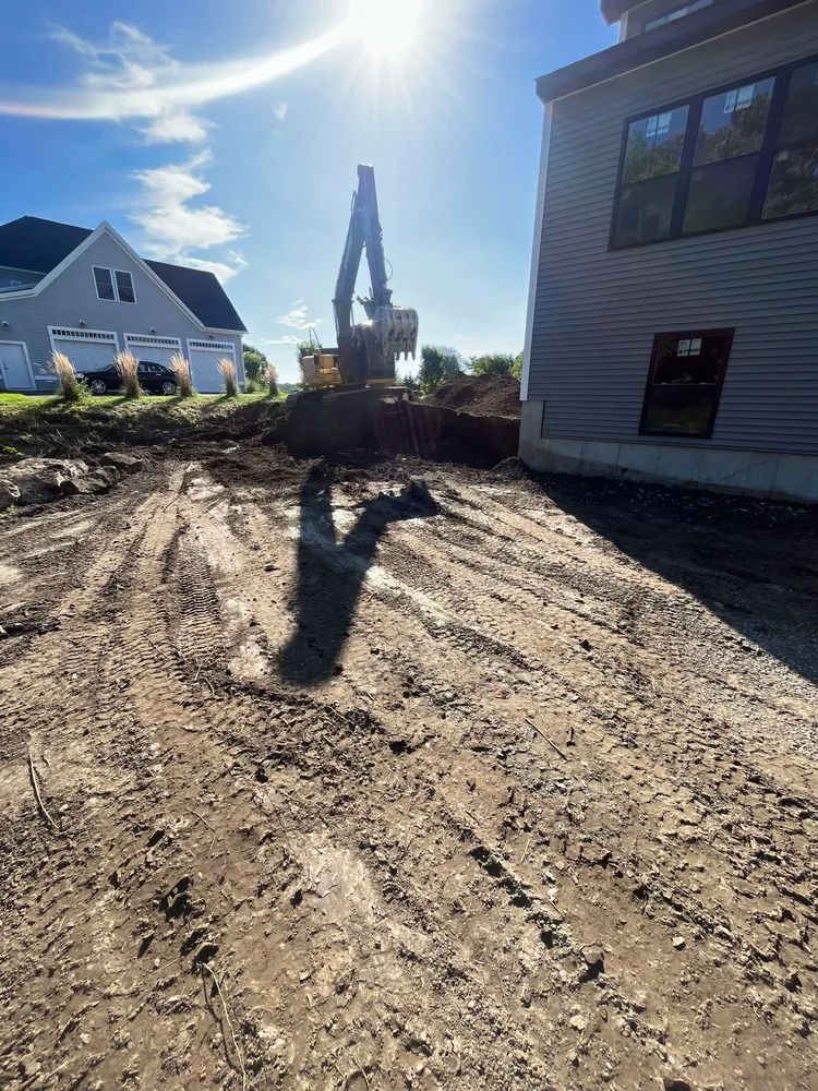 An excavator digging earth near a house under a bright sunny sky. The ground is muddy.