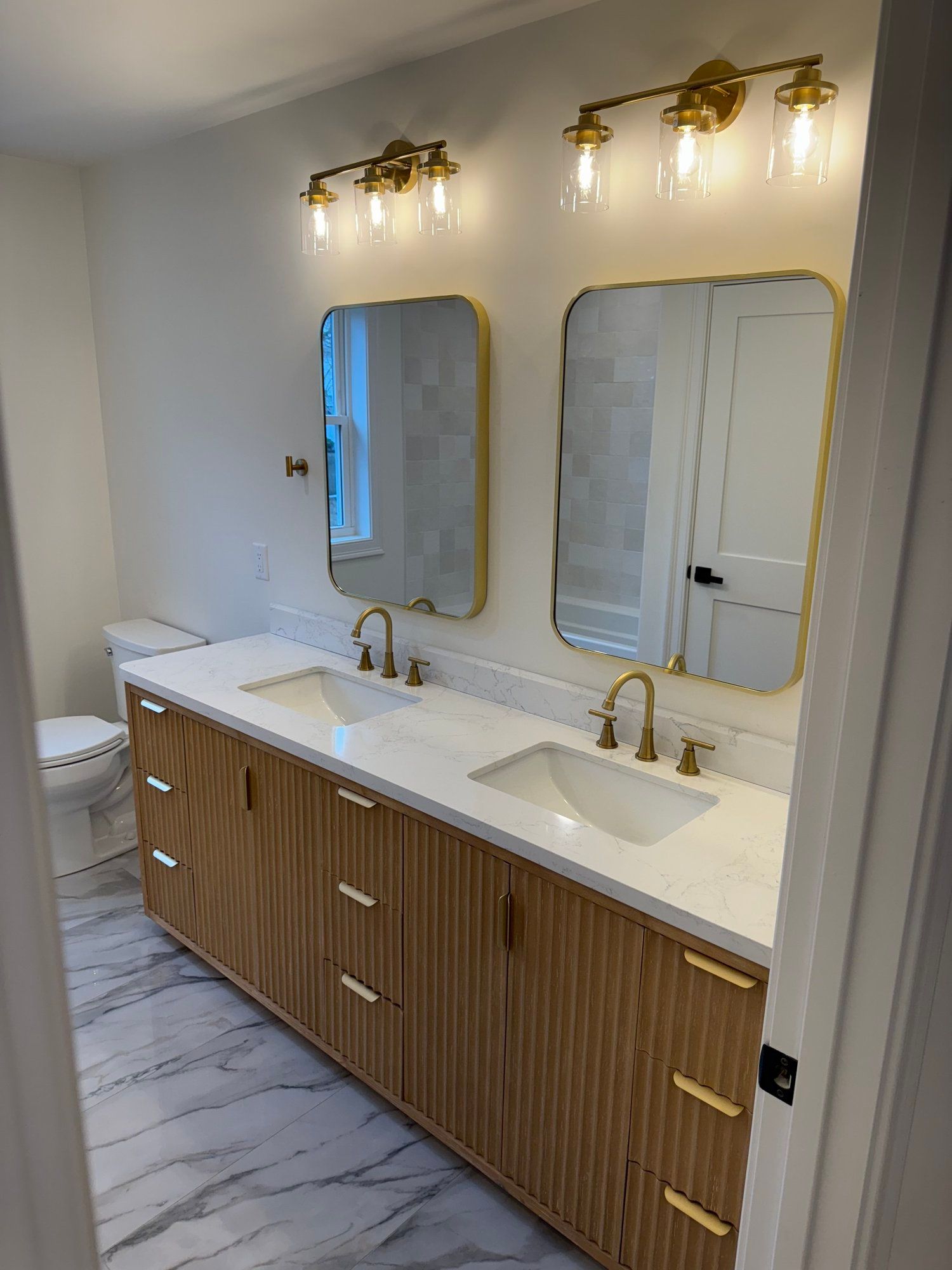 Bathroom with a light wooden vanity, marble countertop, gold accents, and two mirrors.