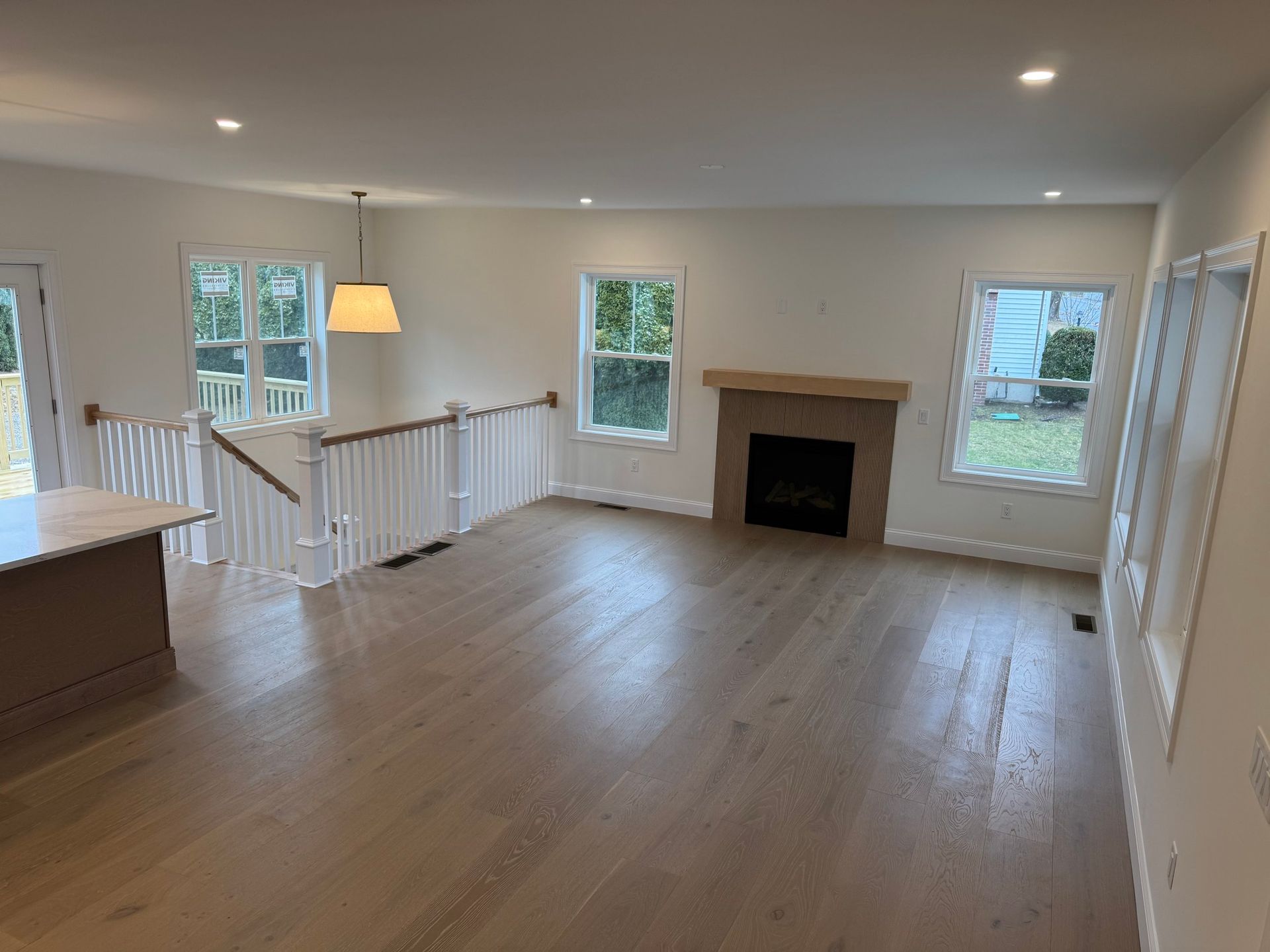 Empty living room with wood floors, fireplace, and staircase. White walls, windows, and light fixture.