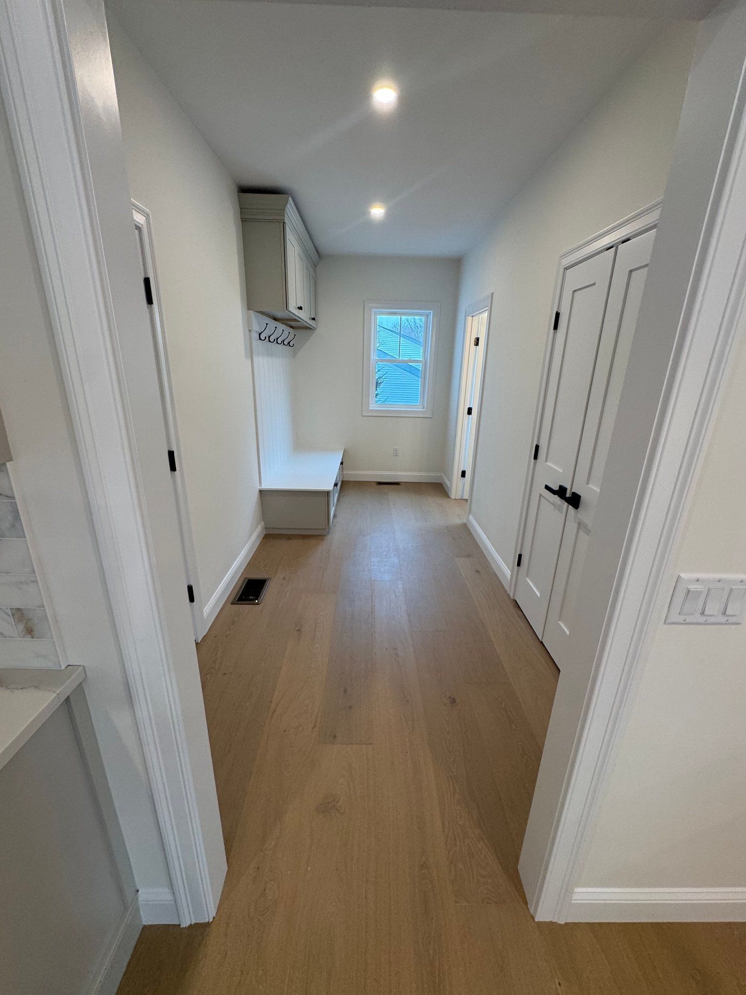 Hallway with light wood floors, white walls, built-in bench, overhead cabinets, and a window at the end.