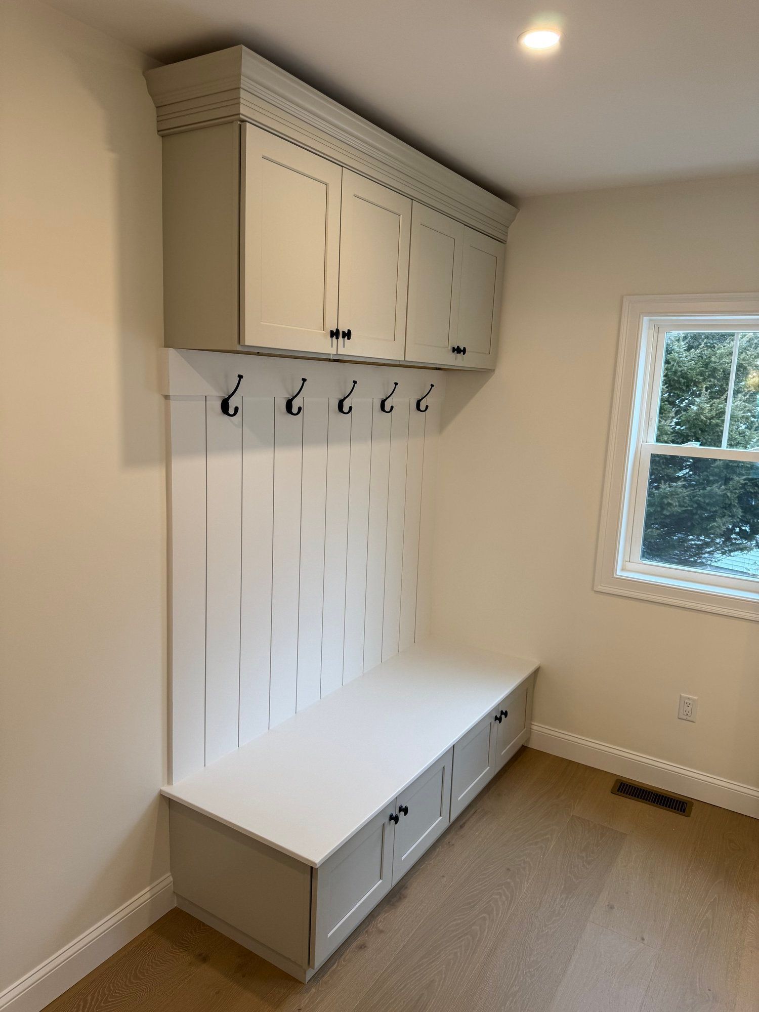 Built-in mudroom storage: gray cabinets and bench, hooks, white paneling, and a small window.