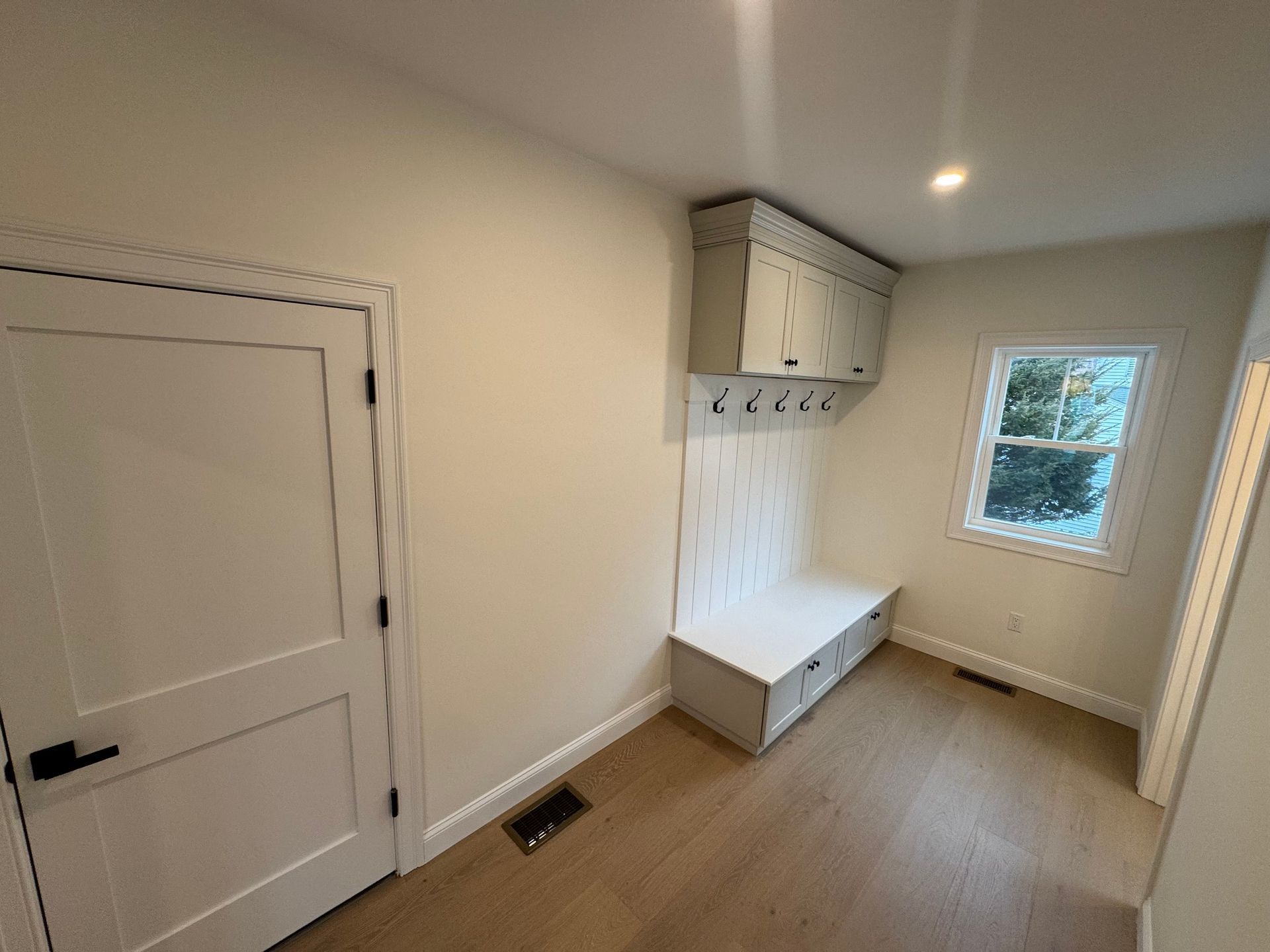 Mudroom with white bench, storage, and hooks. Beige walls, light wood floor, small window.