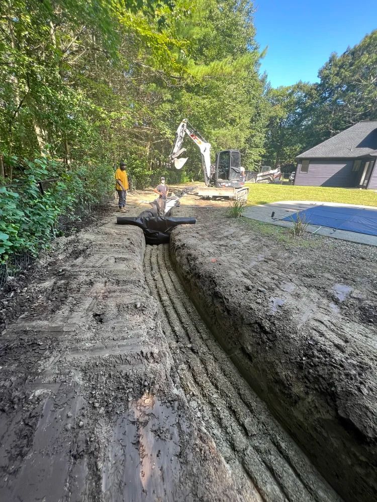 A trench dug in dirt, with buried pipes. A backhoe and people work in the background. Green trees and a house are visible.