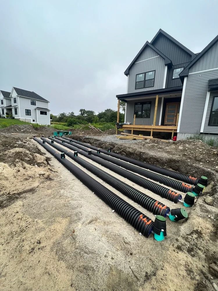 Black drainage pipes laid in trenches near a gray house under construction.