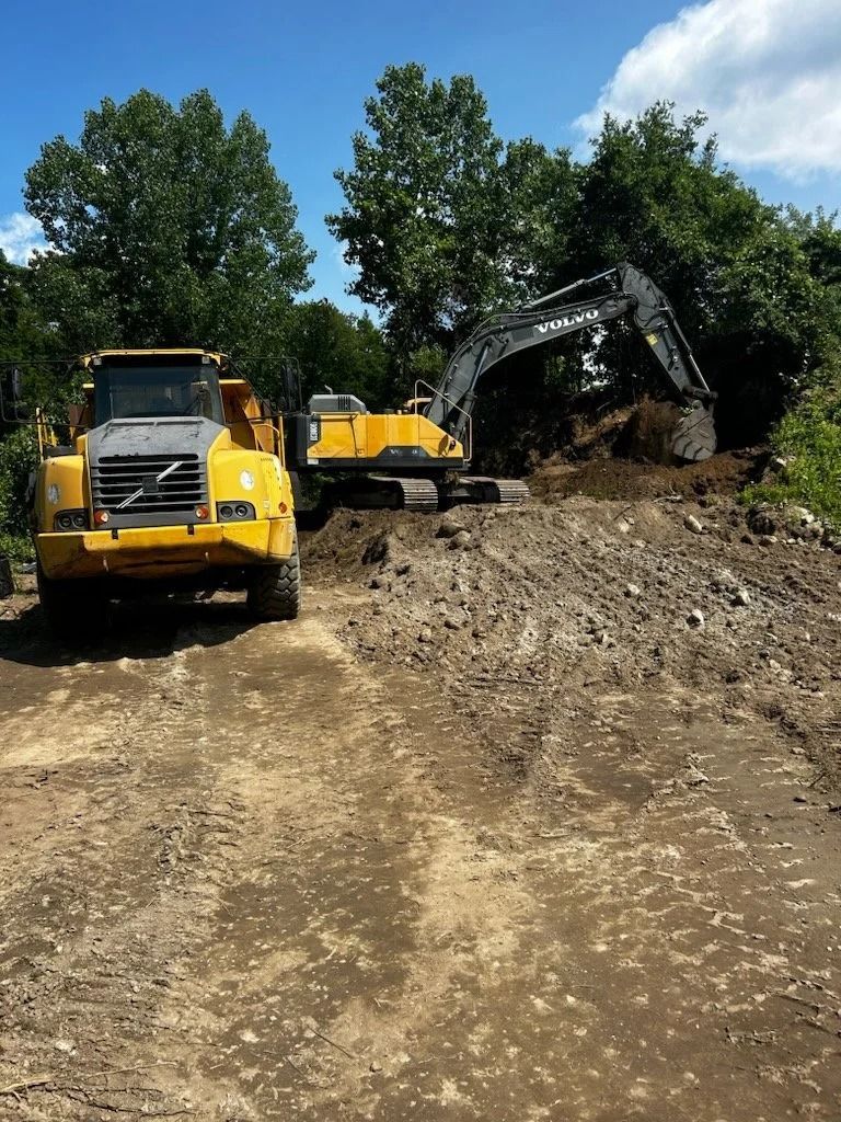 Yellow construction vehicles on dirt road, excavator digging dirt pile. Trees, blue sky in background.