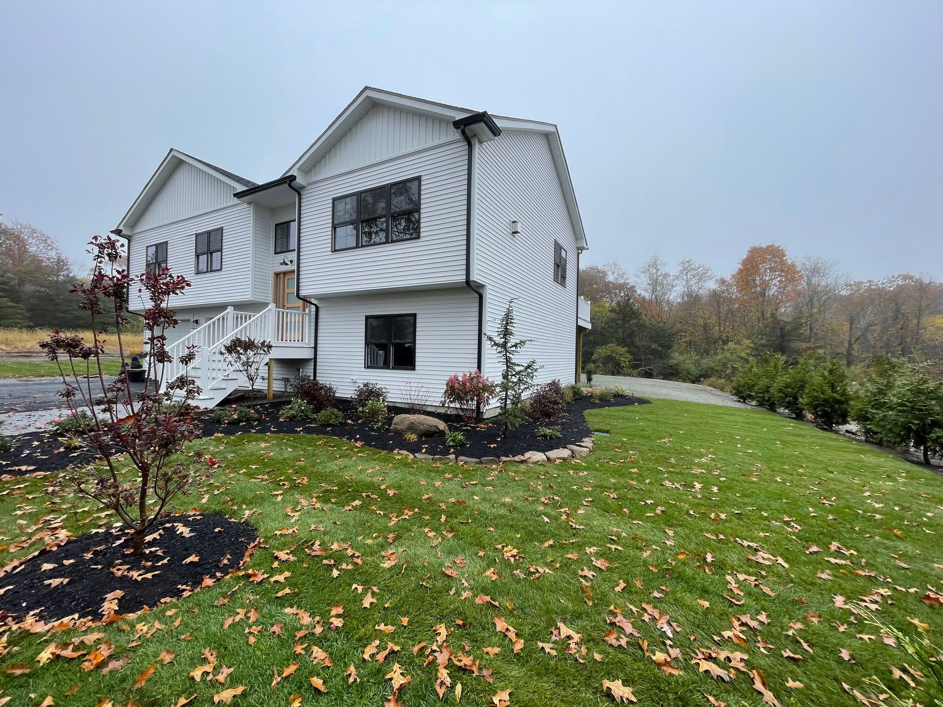 White two-story house with black trim, surrounded by green grass and a gravel driveway on a cloudy day.