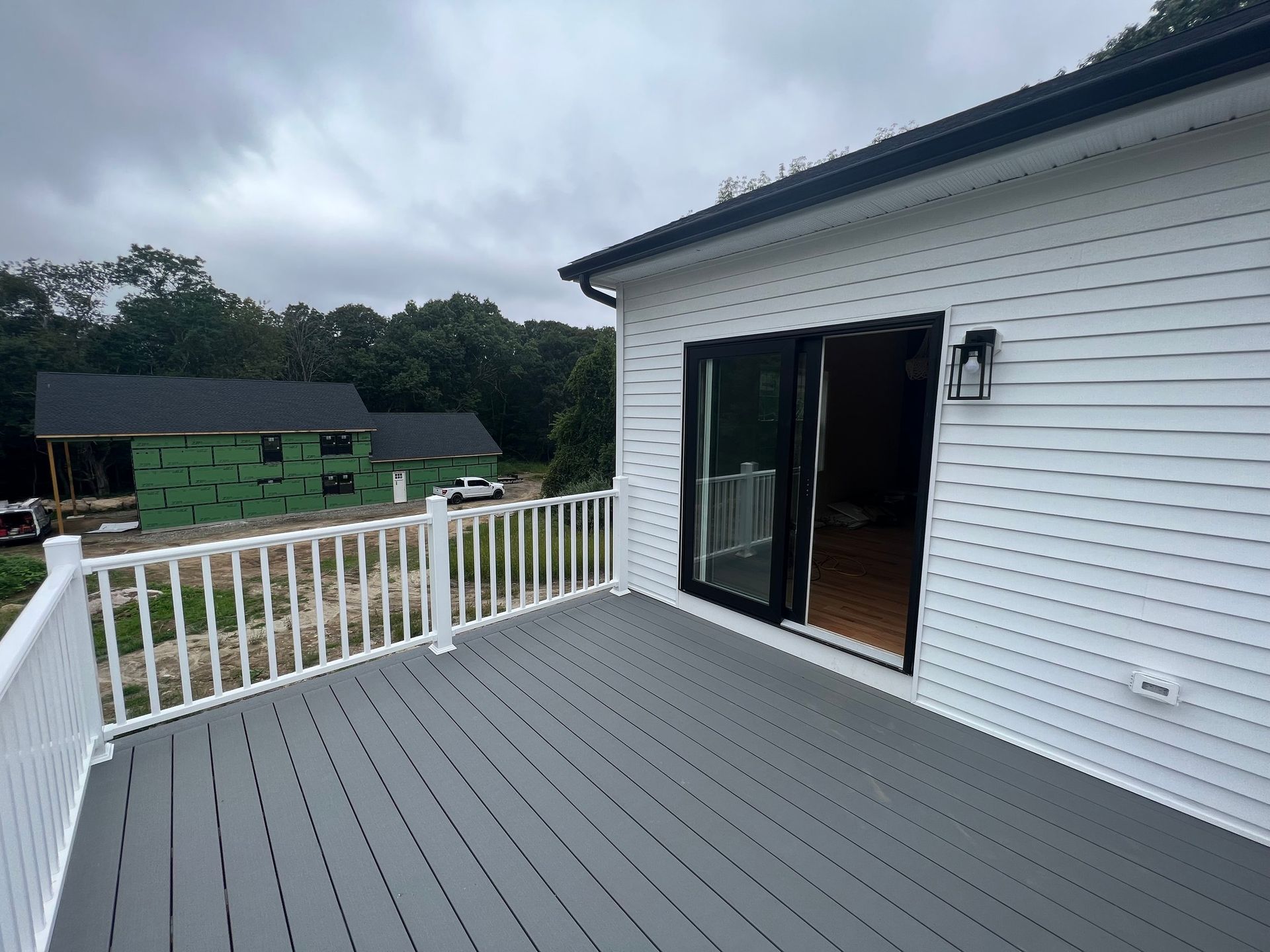 Grey deck with white railing, sliding glass door, and white siding. A partially constructed building is visible in the background.