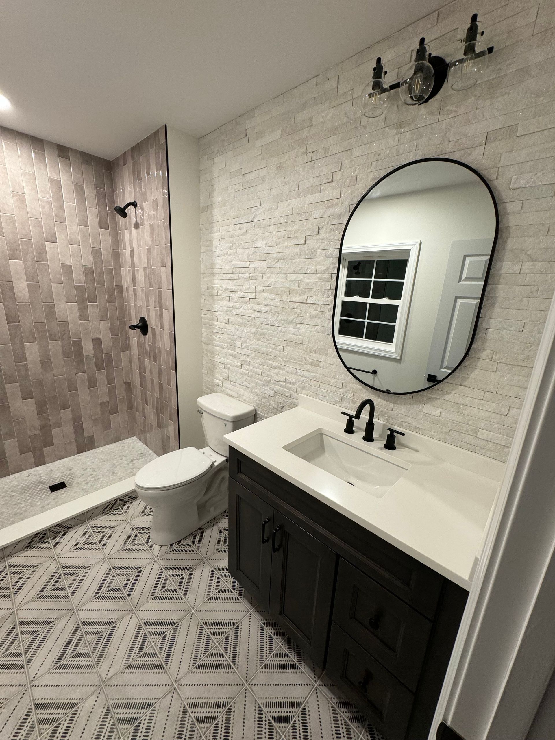 Bathroom with mosaic tile walls, black fixtures, dark wood vanity, oval mirror, and patterned floor tile.