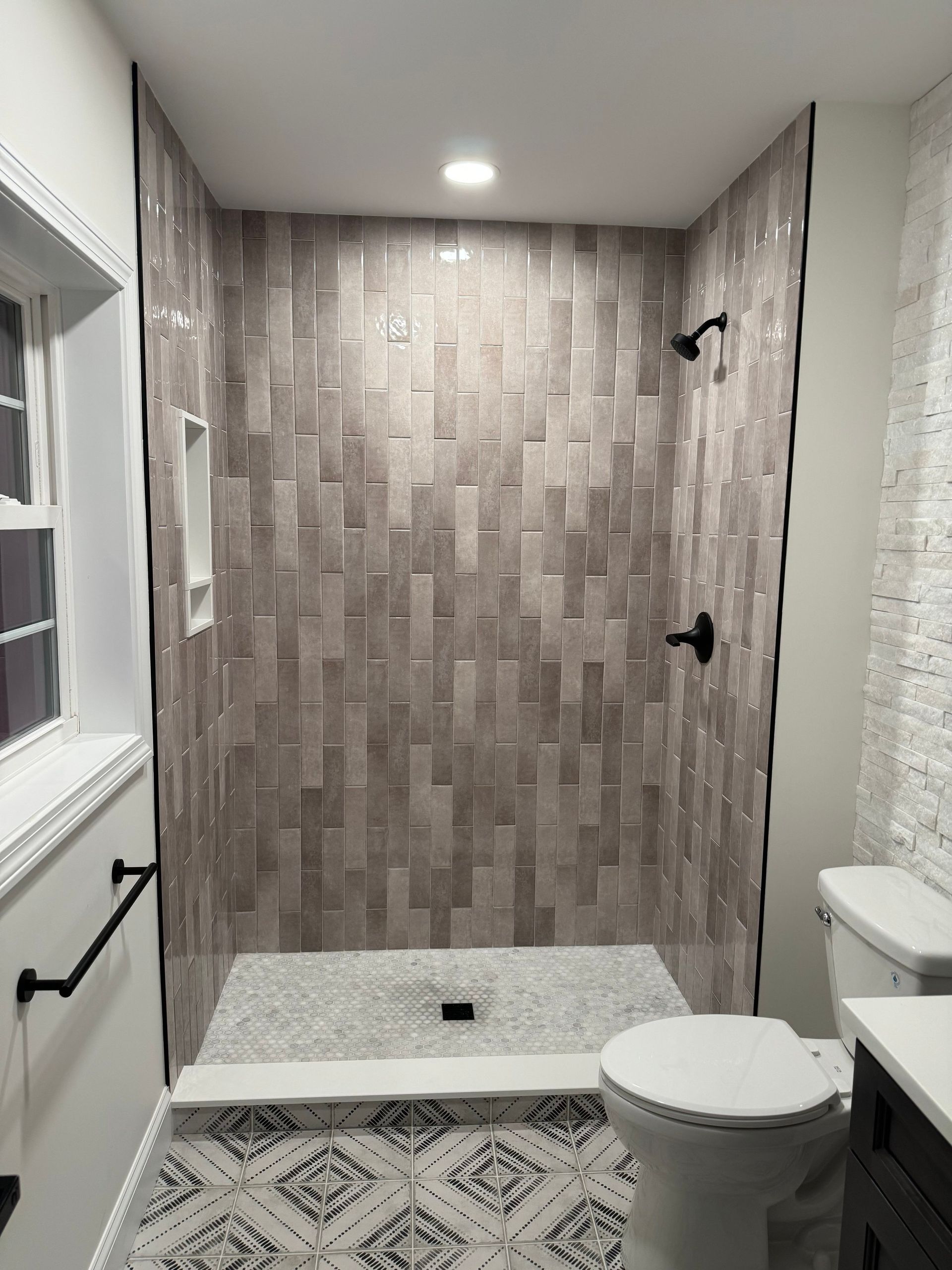 Modern bathroom with tiled shower stall, toilet, patterned floor, and window.