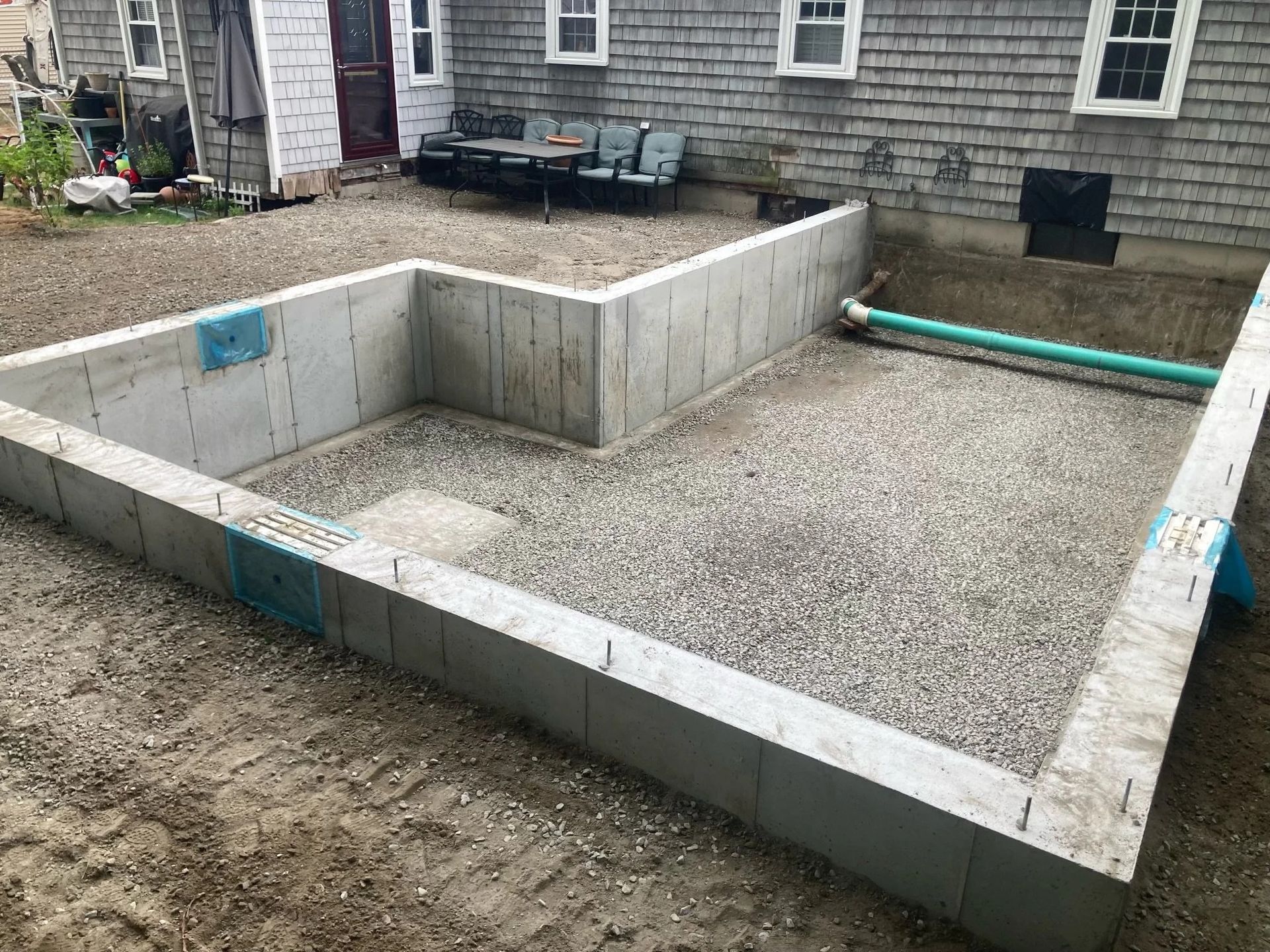 Concrete foundation walls with gravel interior, prepared for a structure. Outdoor setting with house background.
