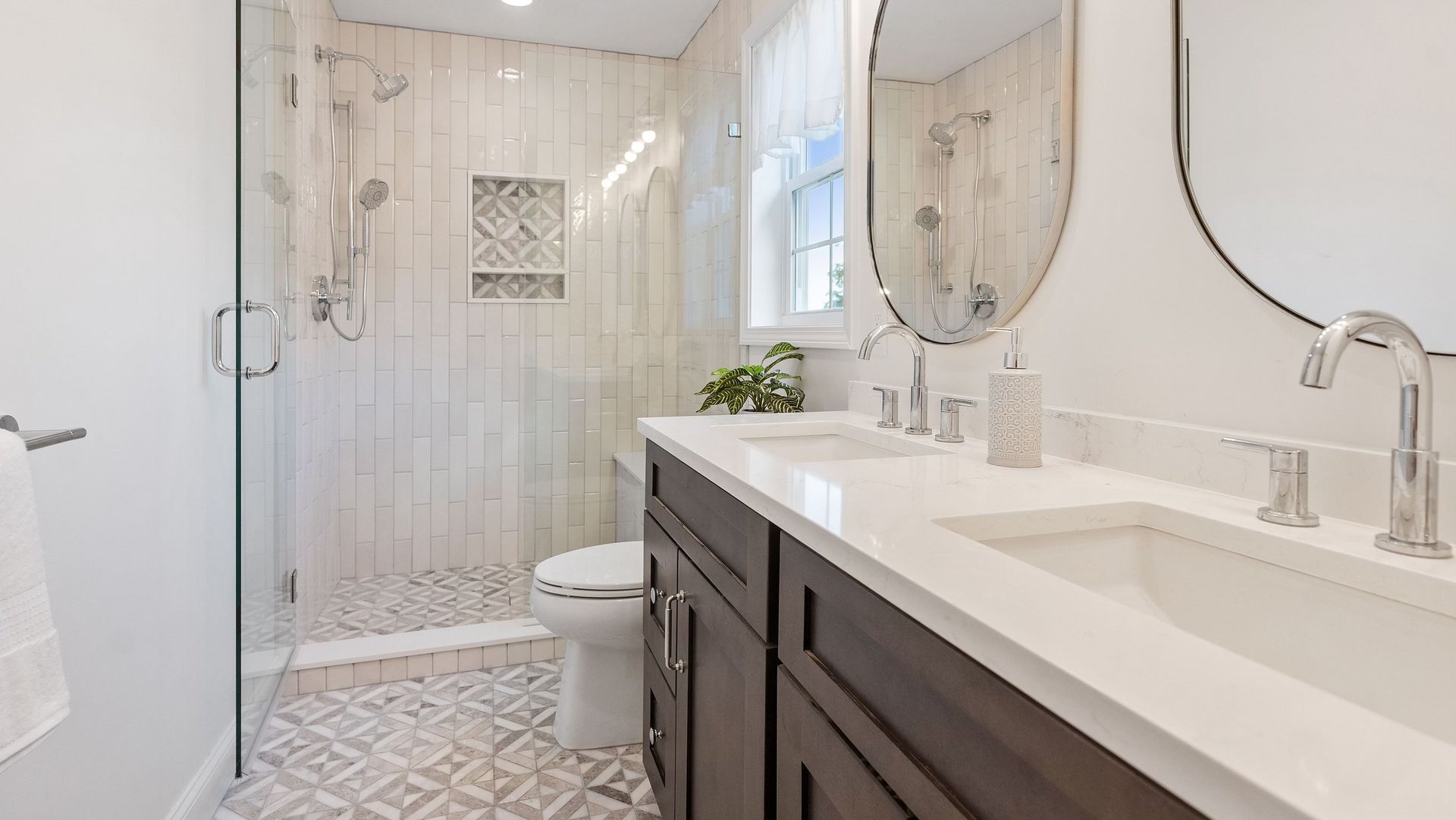 Bathroom with white and gray tile, double vanity, glass shower, and oval mirror.