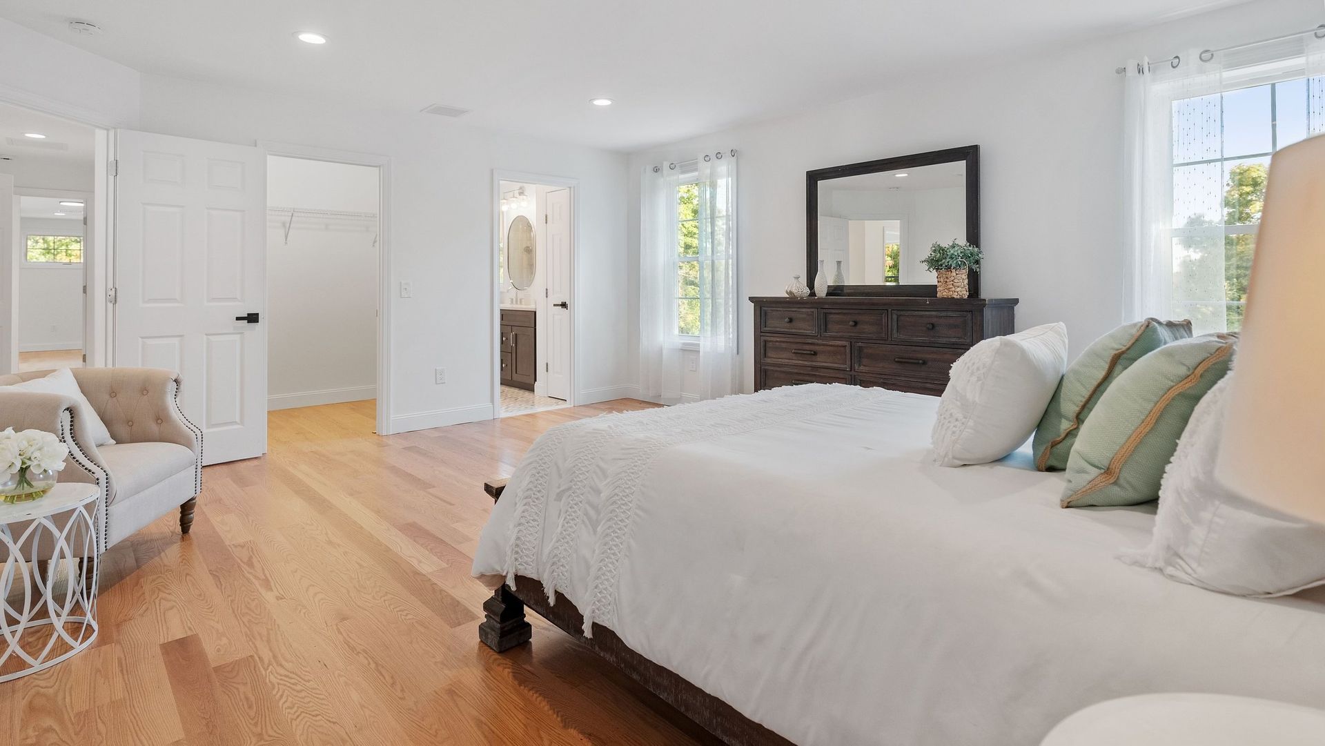 Spacious bedroom with hardwood floors, a large bed, dark dresser, and windows with sheer curtains.