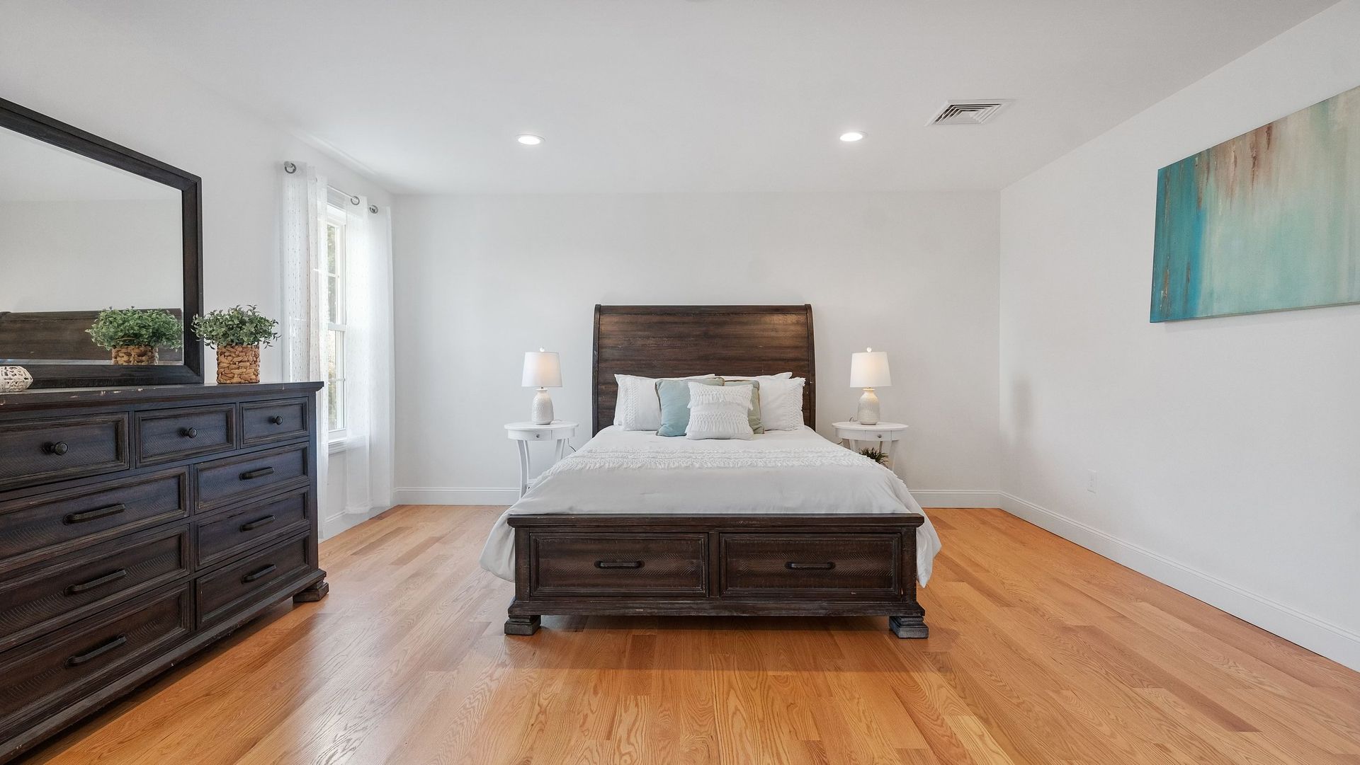 Bedroom with wooden bed, dresser, and hardwood floors. White walls and artwork.