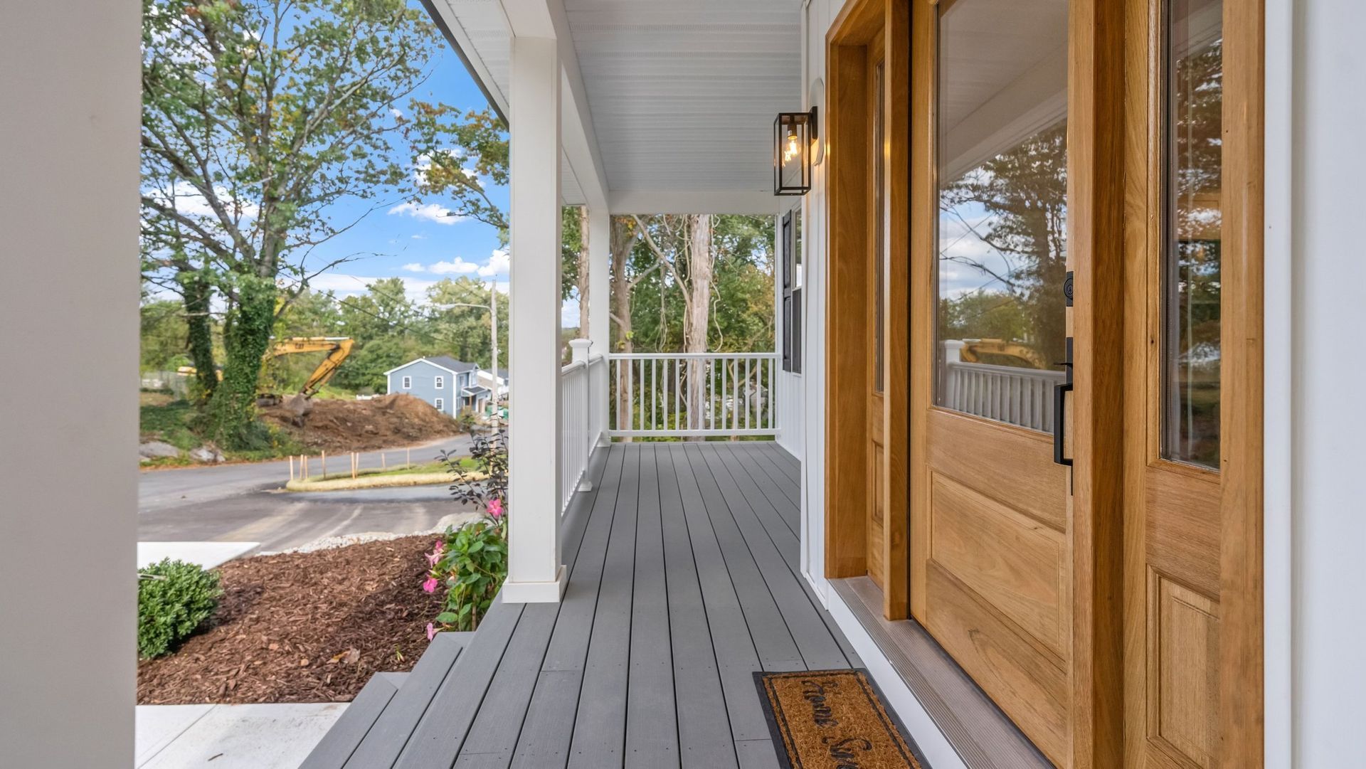 Gray porch with wooden door, white columns and railing. Brown mulch and greenery on the left.