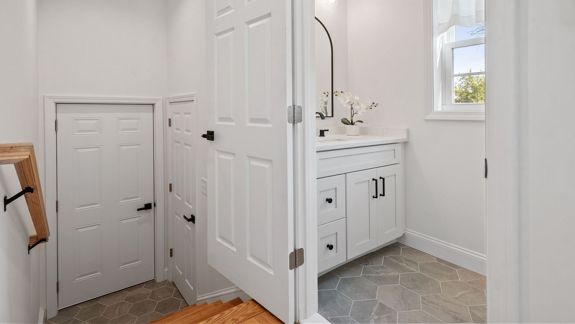 White bathroom with a door and a small storage door, white cabinets and a window.