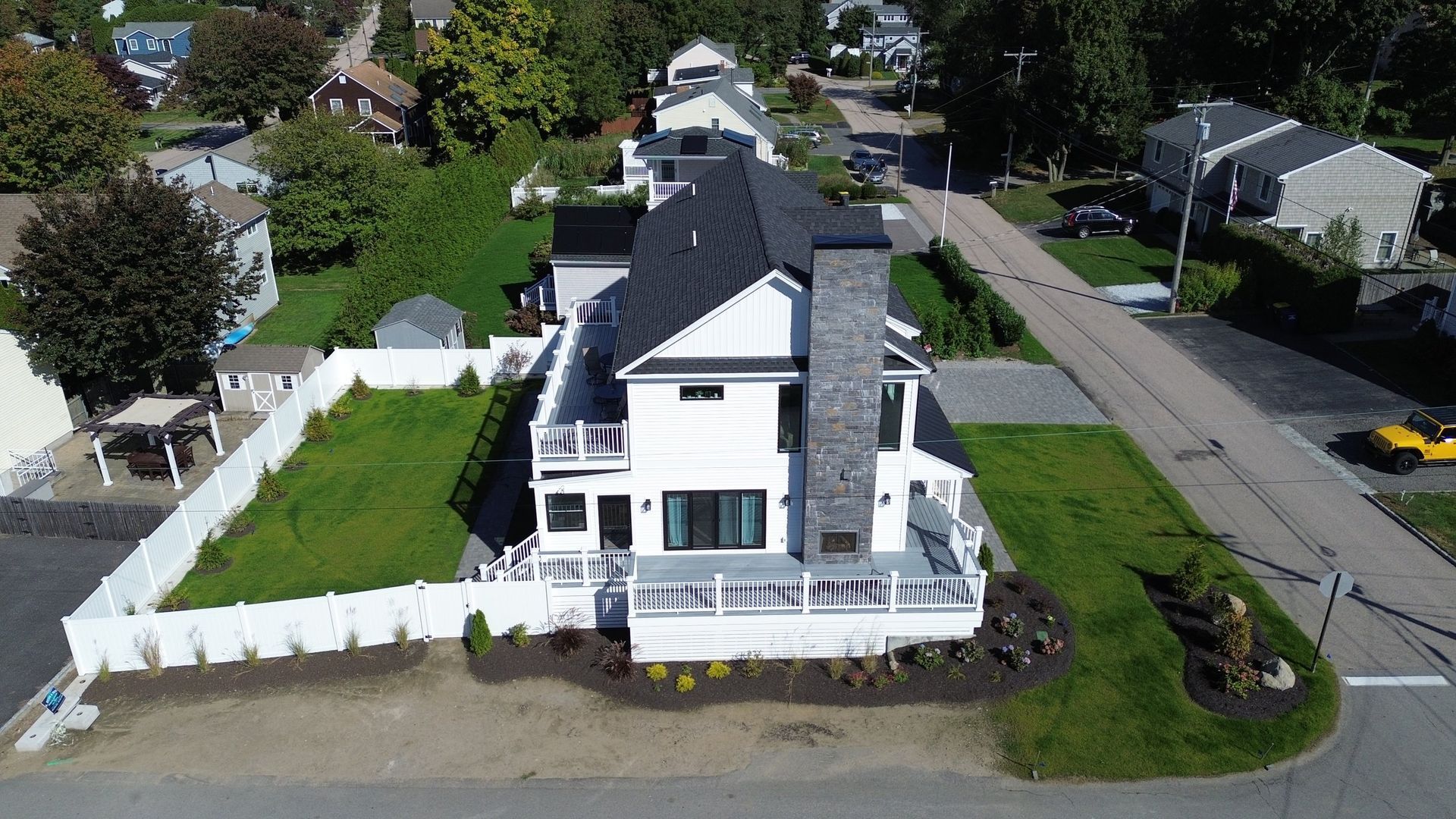 Aerial view of a white house with a dark roof and a stone chimney, surrounded by green grass and a white fence.