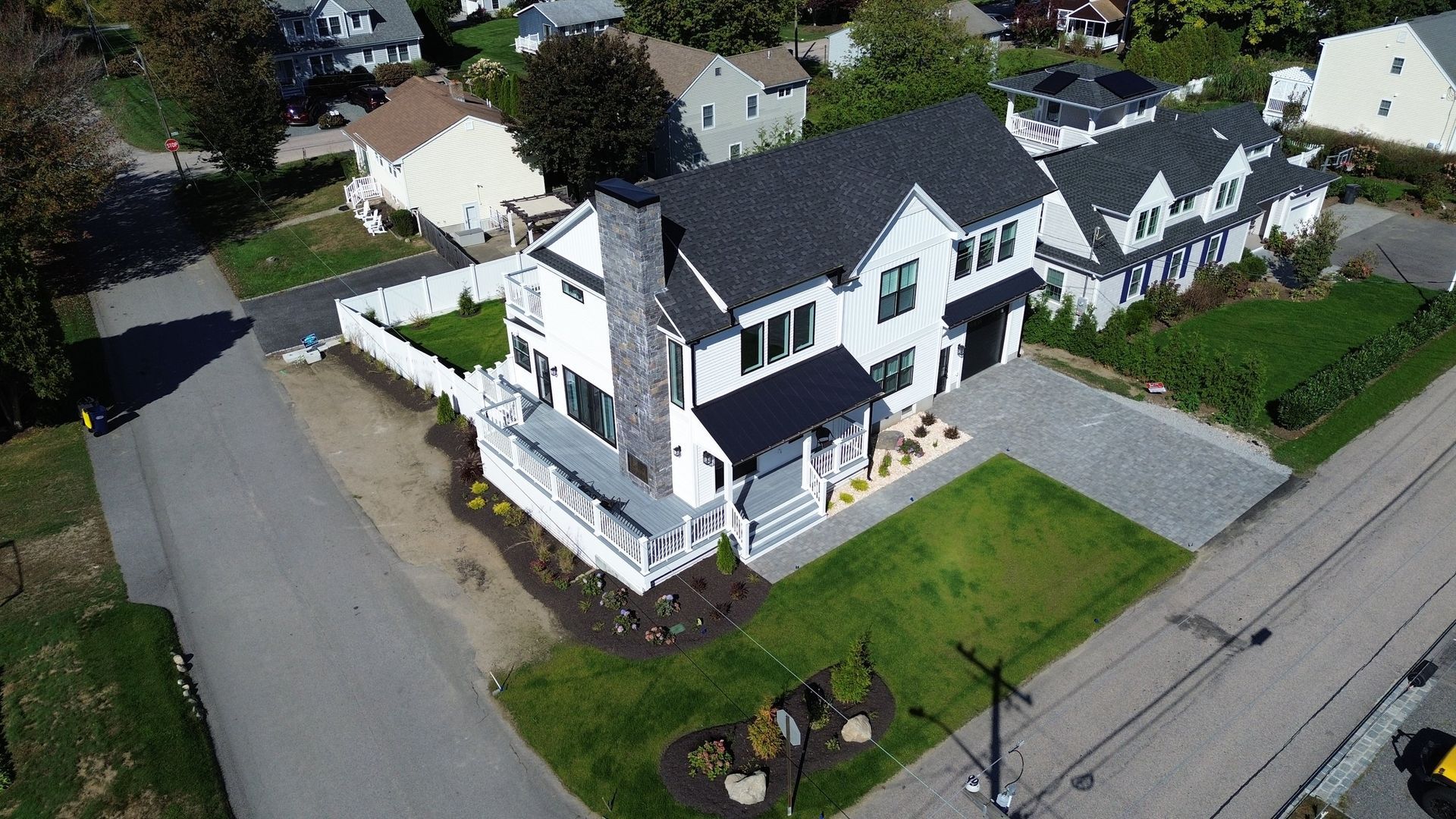 Aerial view of a white house with black roof, chimney, and trim; on a green lawn with a gray driveway.