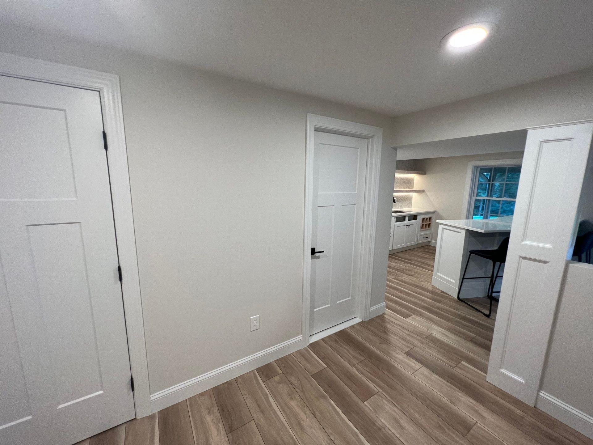 Interior view of a room with wood-look flooring, white doors, and a partial view of a kitchen area with a window.