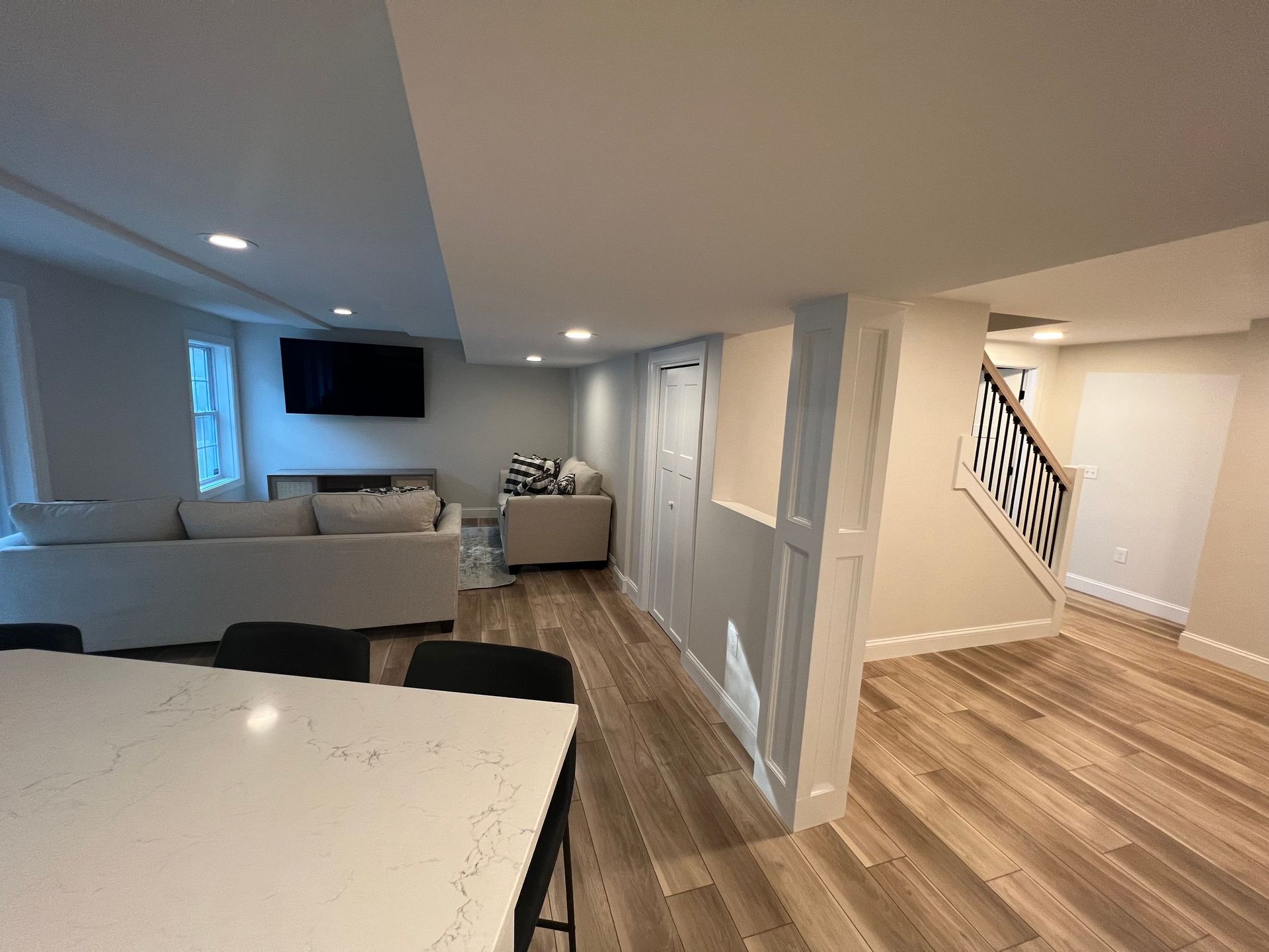 Modern open-plan basement with living area, kitchen island, and stairs leading upwards, lit by recessed lighting.
