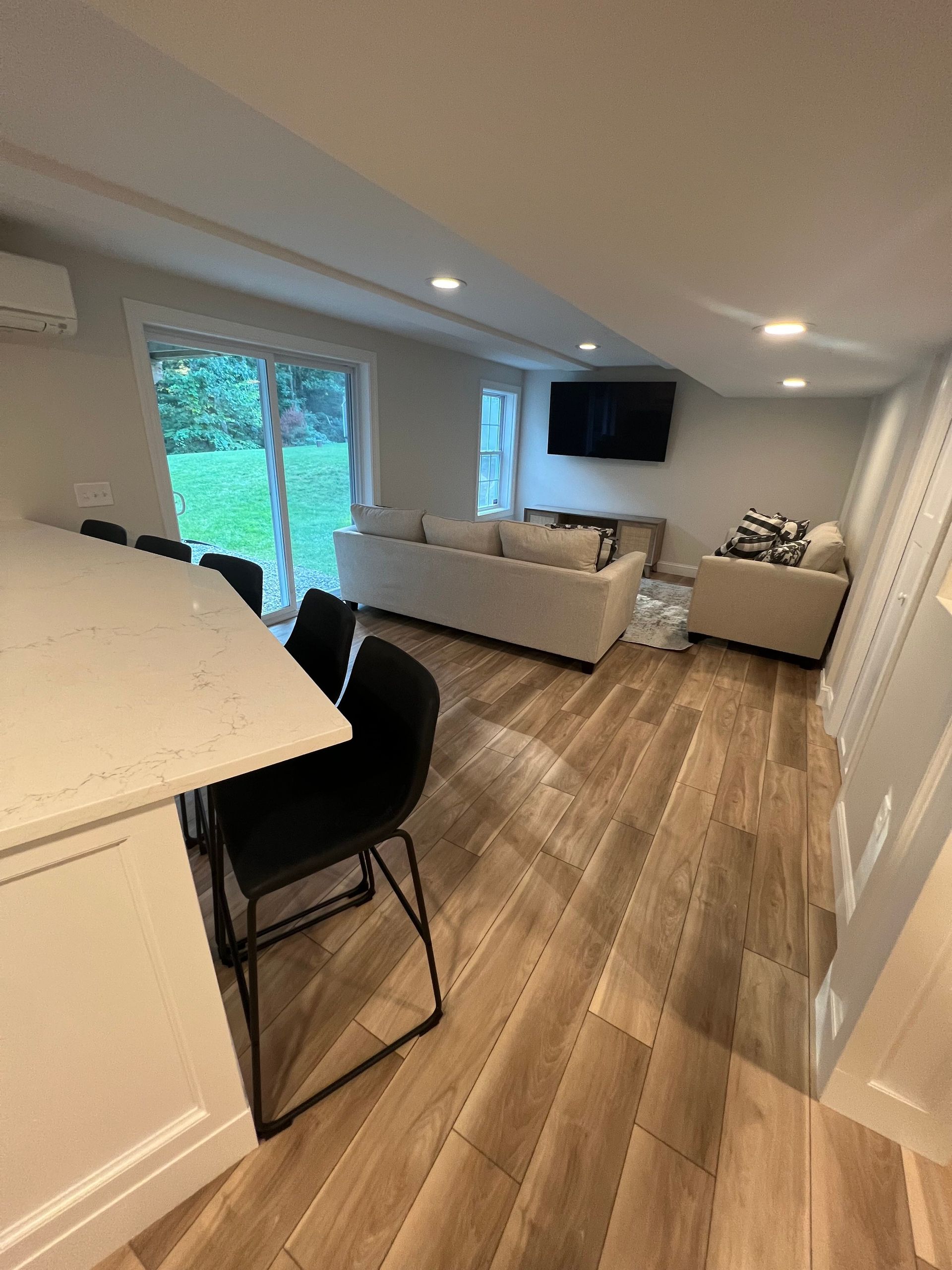 Modern living room with light-colored sofa, TV, and breakfast bar. Sliding glass door overlooks a grassy area.