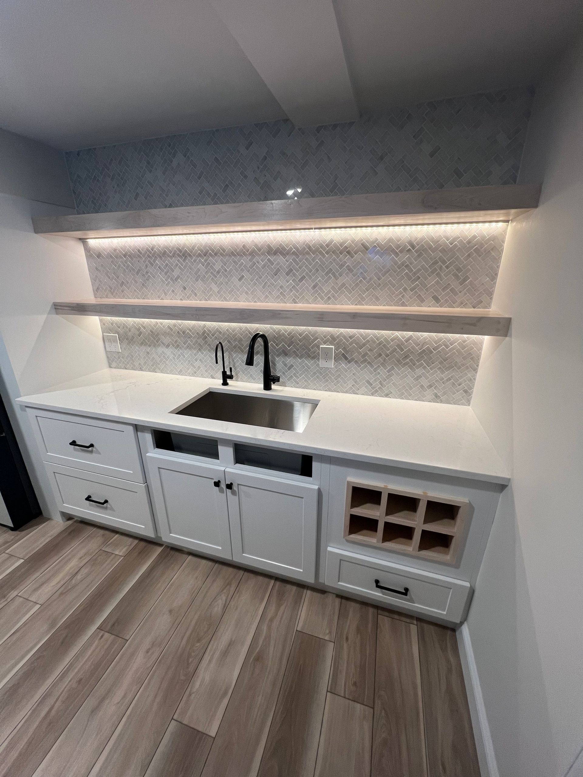 White bar cabinet with sink, black faucet, wine rack, and under-cabinet lighting against a mosaic backsplash.