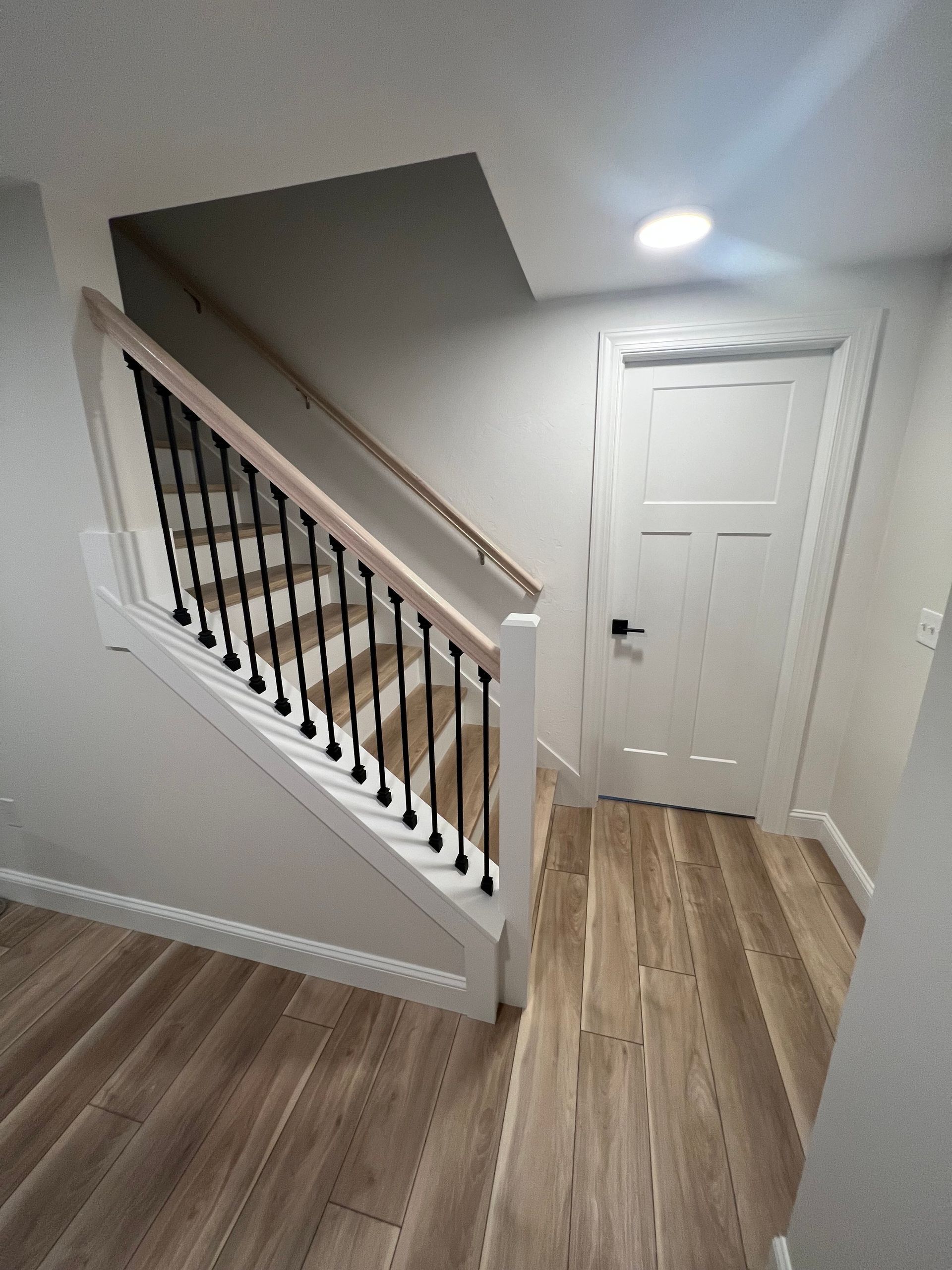 Staircase with black spindles, light wood railing, and a white door with wood-look flooring.