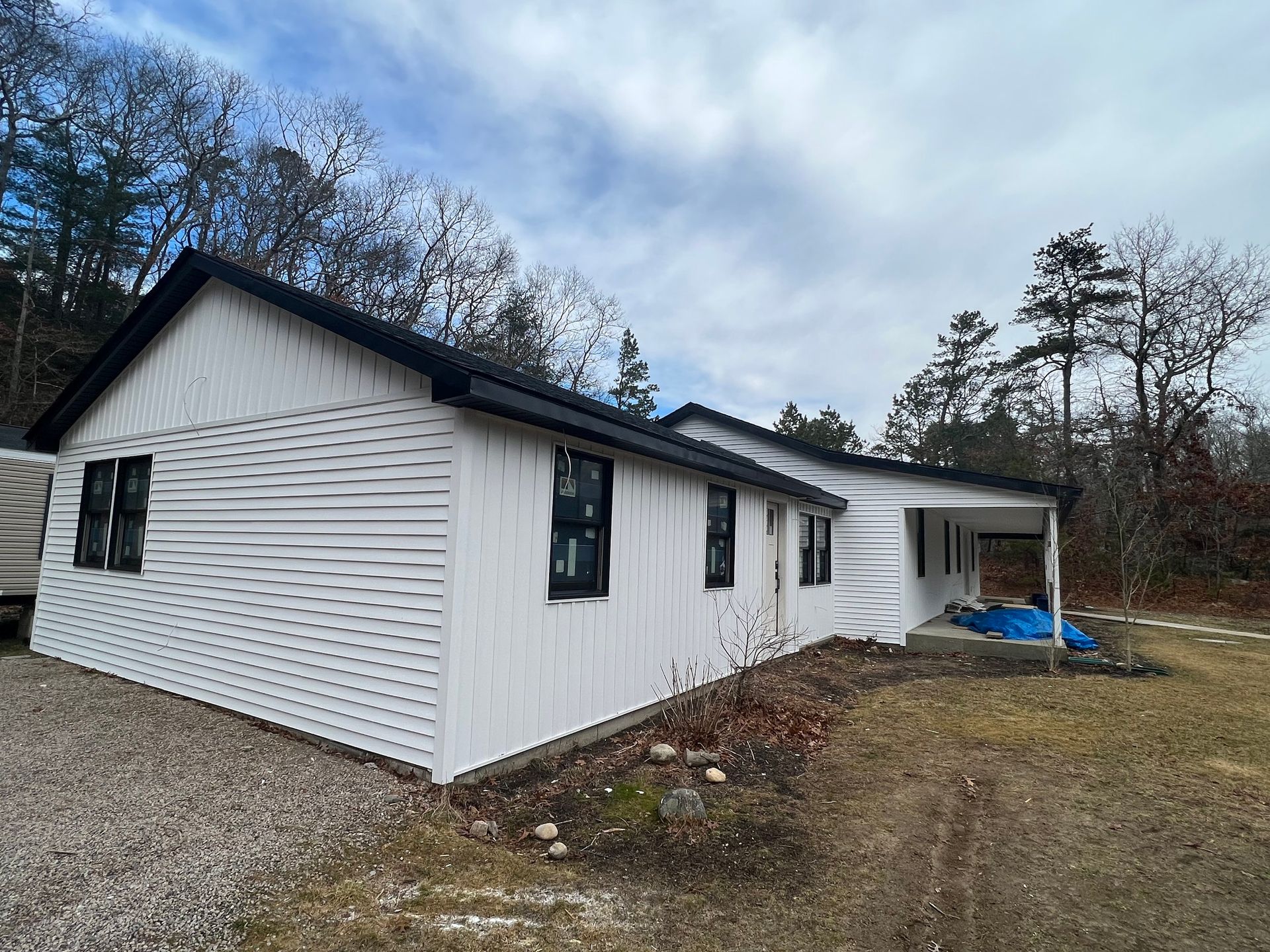 White house with black trim, windows, and roof; cloudy sky, trees, gravel yard.