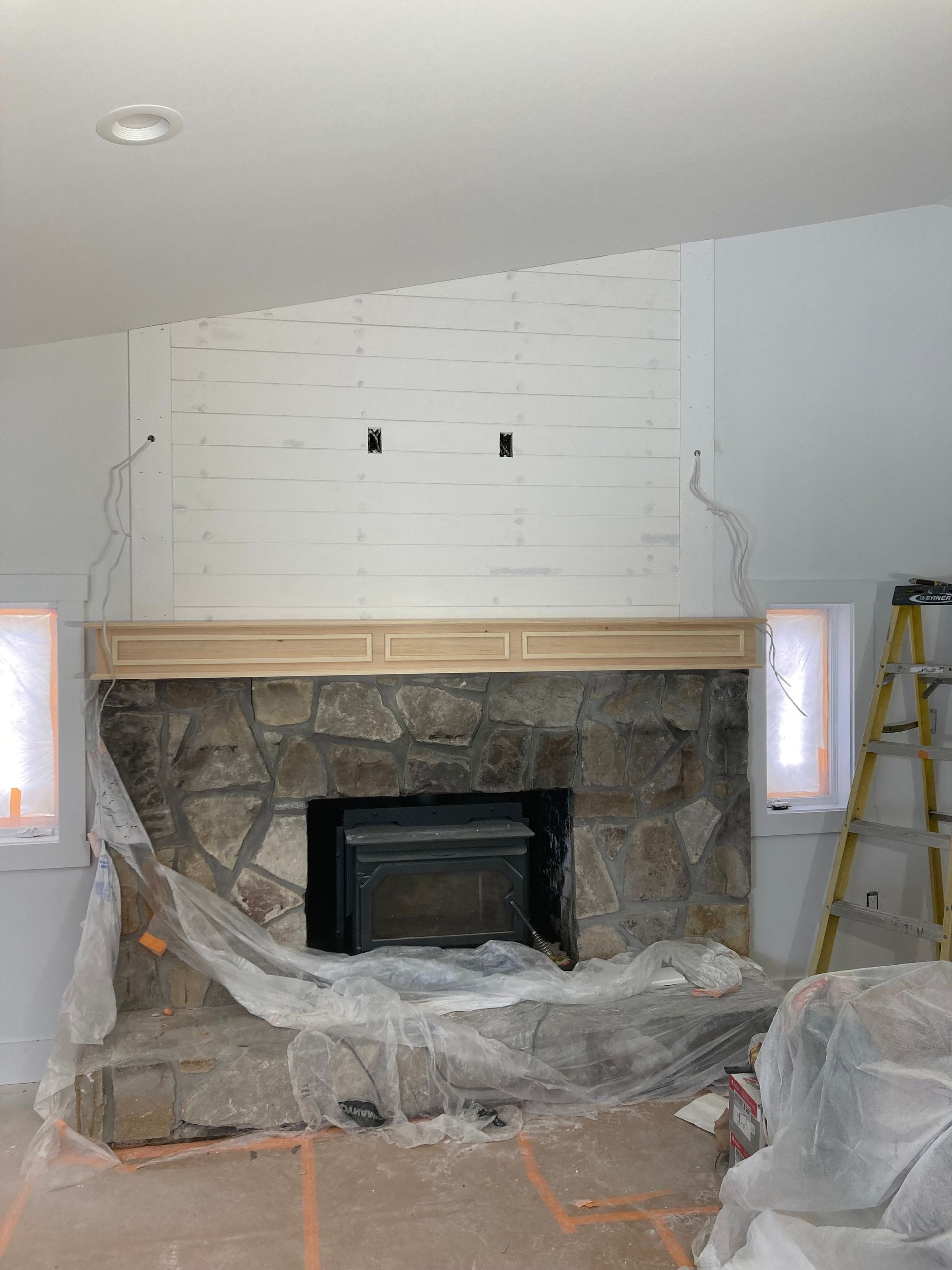 Fireplace with stone surround, wooden mantel, and partially finished shiplap wall. Wiring visible.