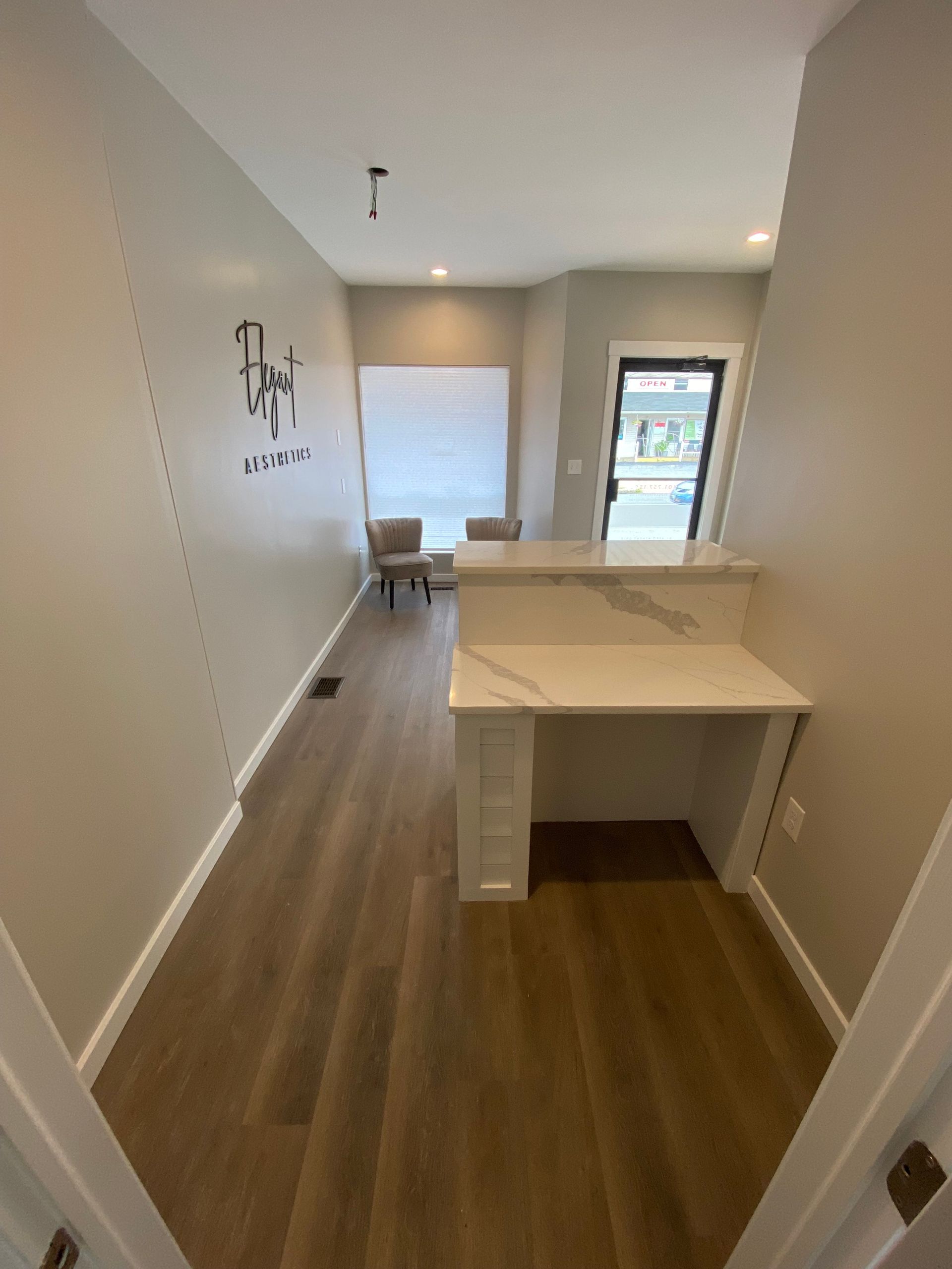 Narrow hallway with wood-look flooring, built-in desk, and doorway leading to a brighter room with chairs and a window.