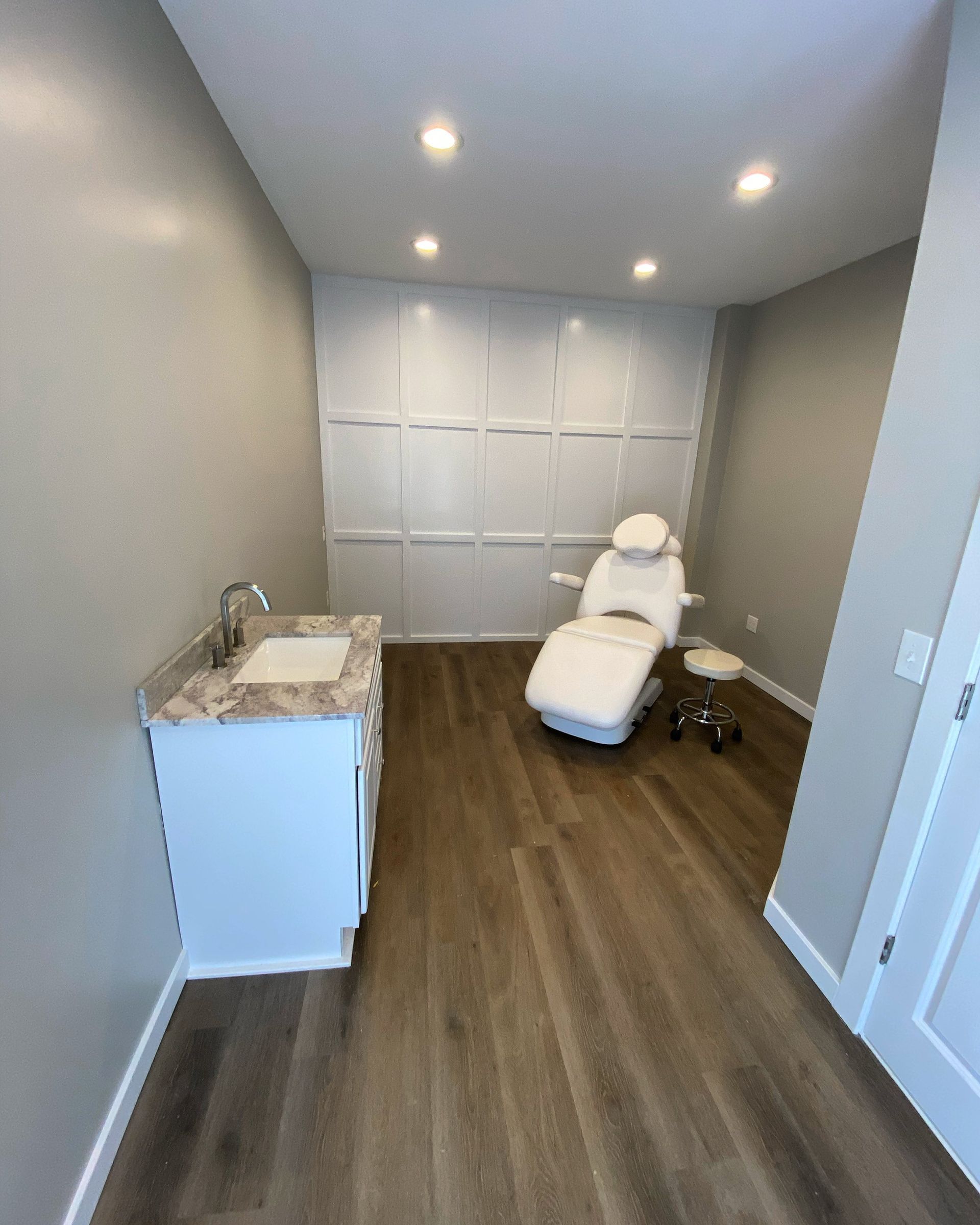 A treatment room with a white chair, sink, and panelled wall. Wooden floor.