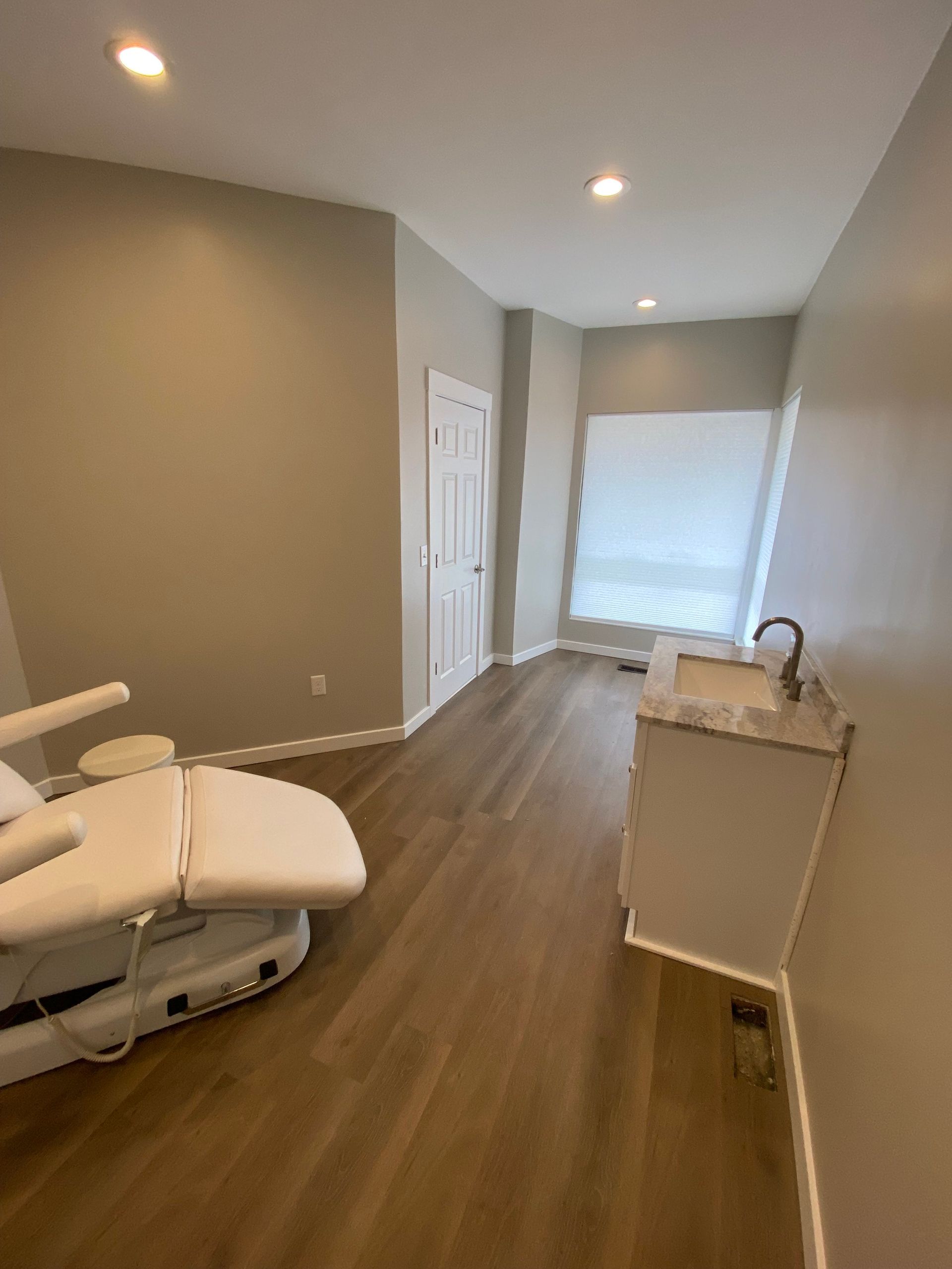 A medical exam room with a reclining chair, sink, white door, and a window with a shade.