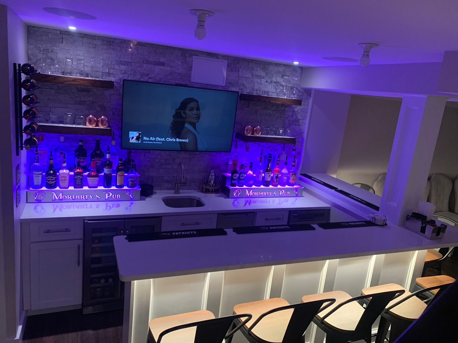 A well-lit home bar with a white countertop and seating, liquor bottles, and a TV. Lit with blue and white lights.
