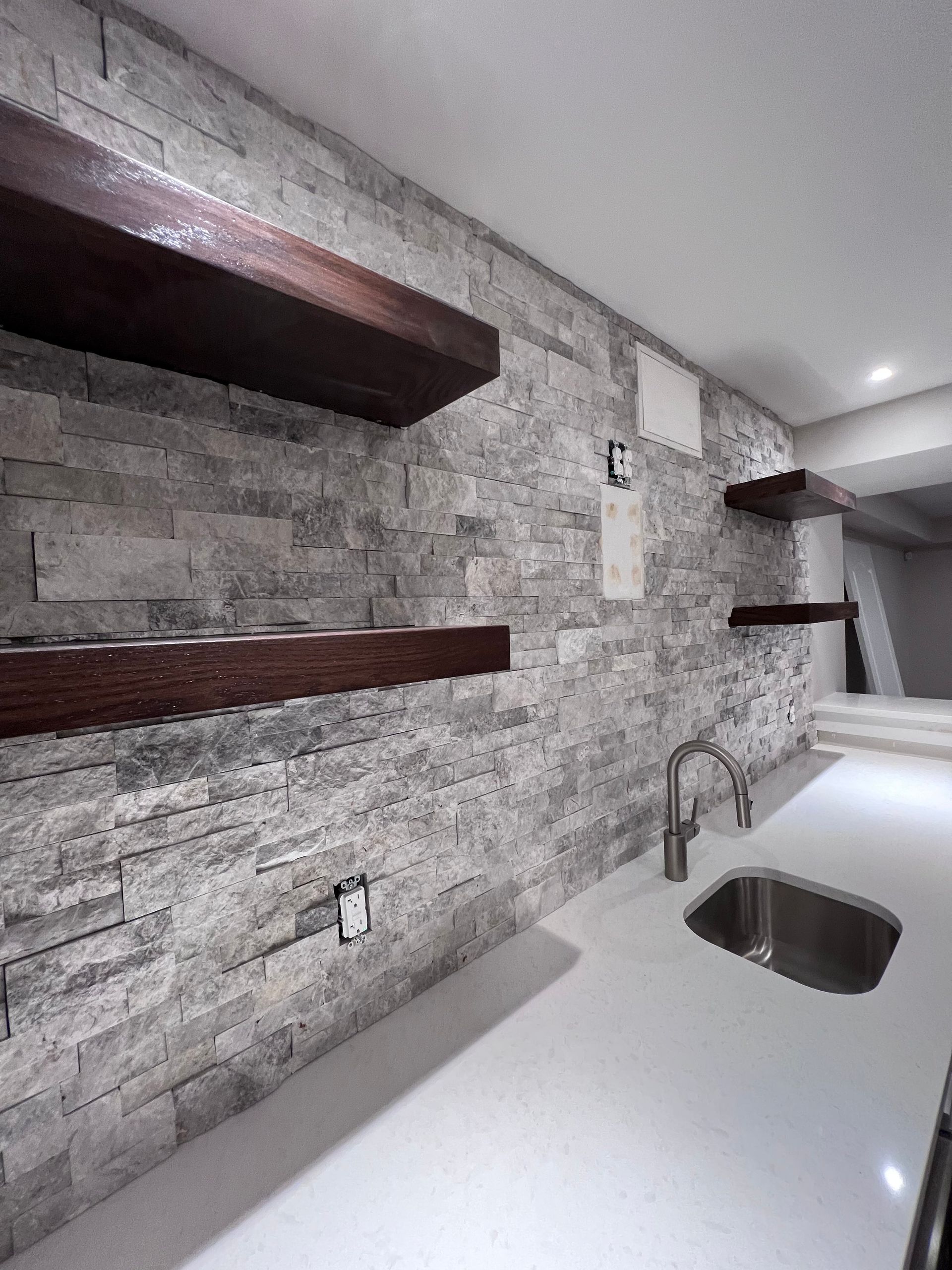 Stone wall with floating wooden shelves above a white countertop and stainless steel sink.