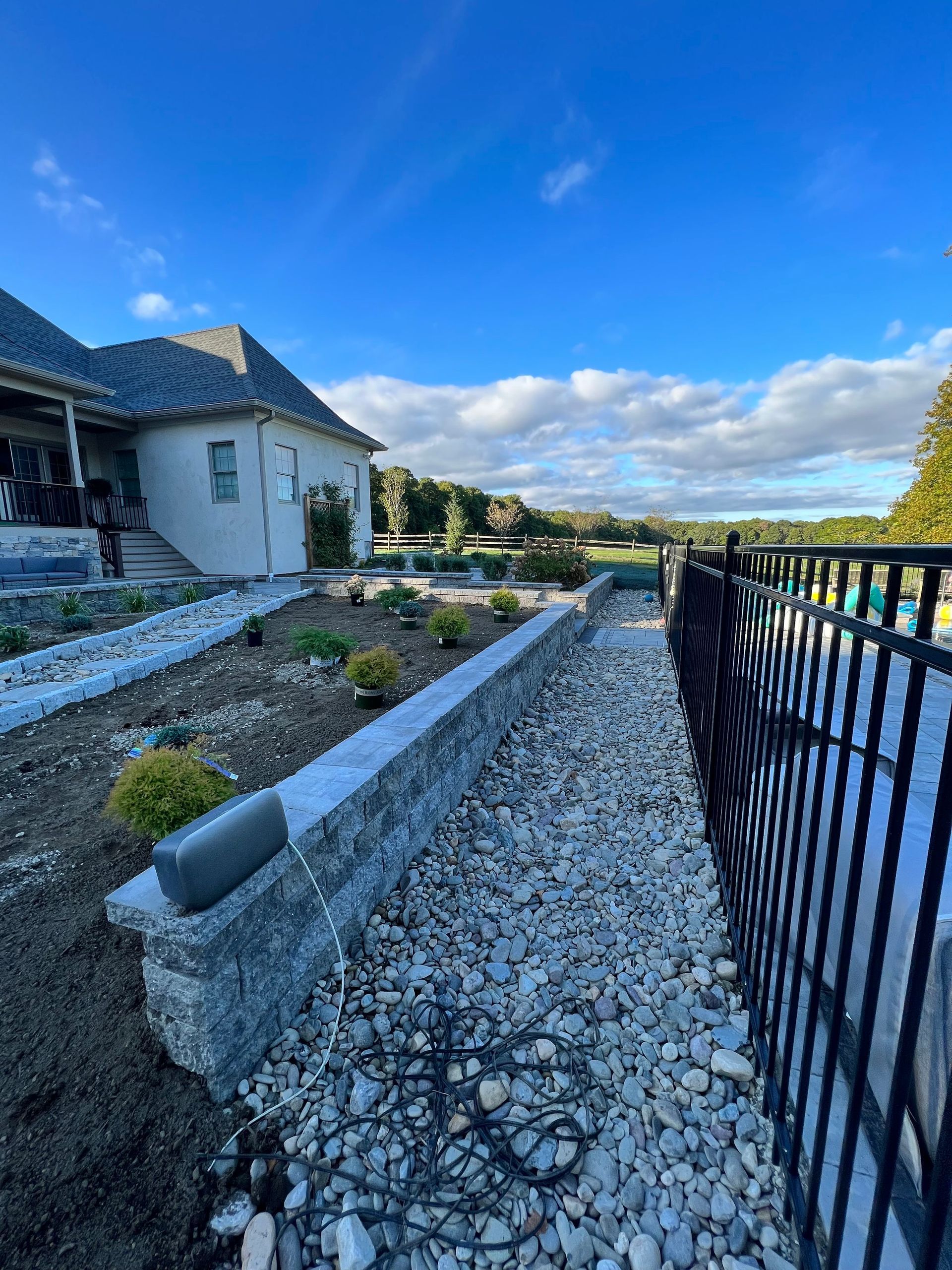 Stone retaining wall and gravel path alongside a black fence and house with blue sky.