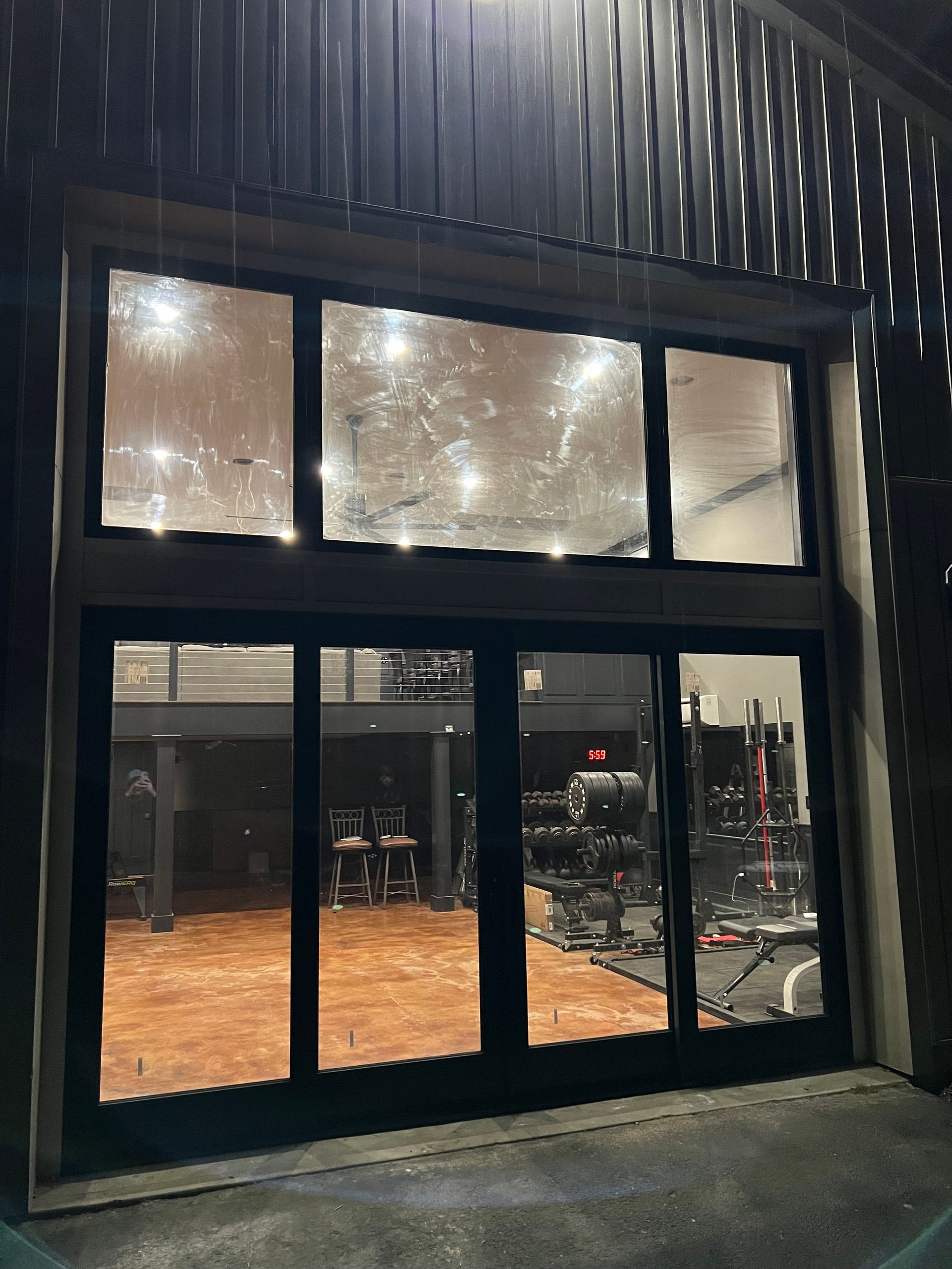 Exterior view of a gym with large windows. Equipment is visible inside, wood floors and black accents. It is nighttime.