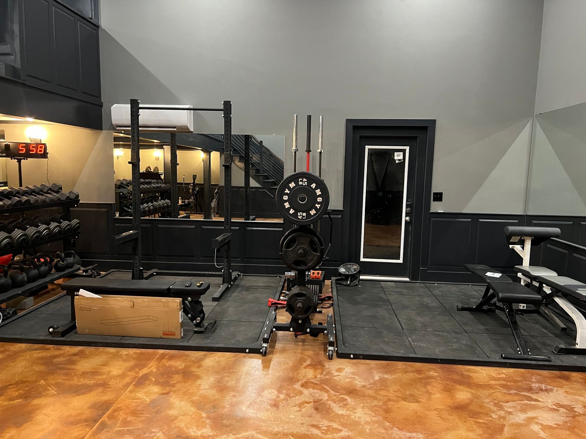 Home gym with weights, rack, and benches on rubber mats, against a dark gray wall and door.