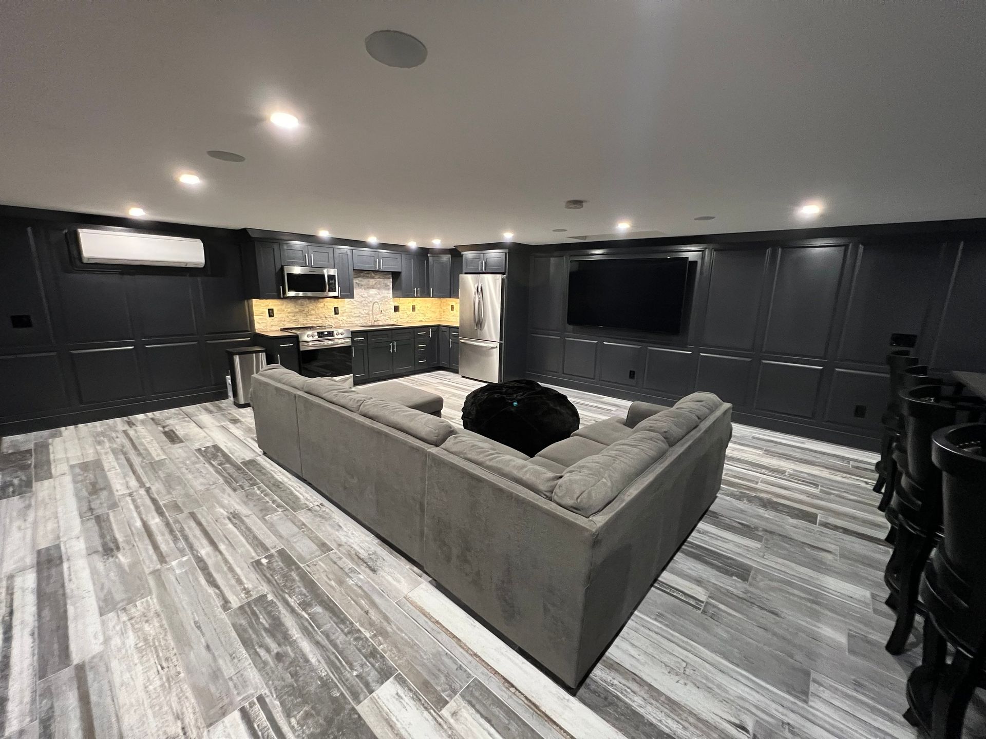 A modern basement lounge with gray sectional sofa, wet bar, and large TV. Dark walls and gray wood-look flooring.