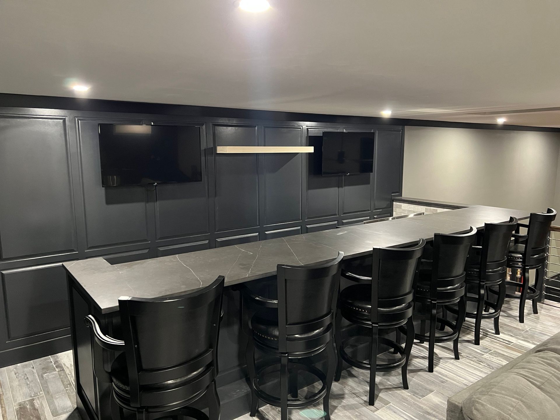 A black bar with stools, two TVs on the wall, and recessed lighting in a finished basement.