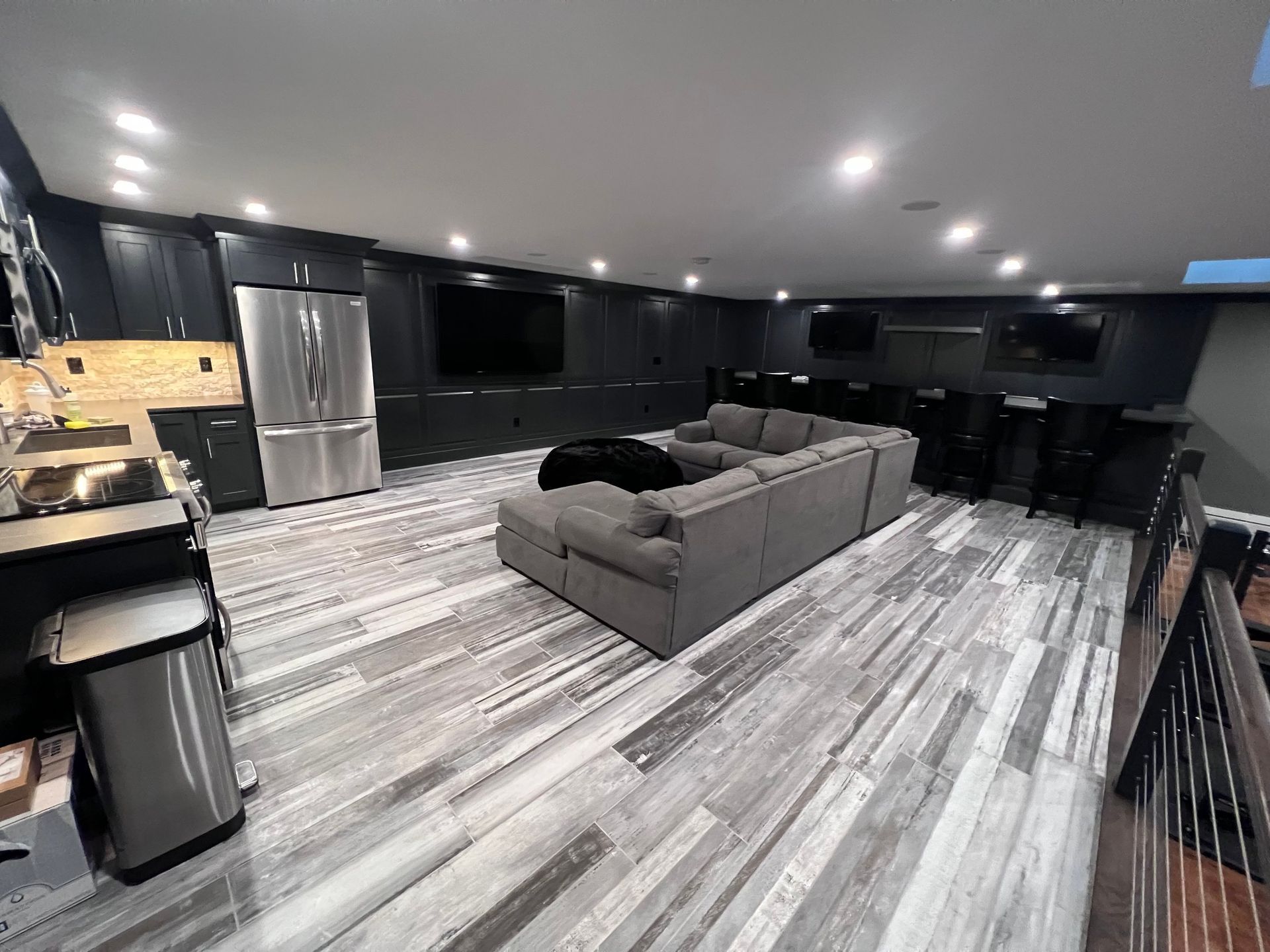 Modern basement with gray wood-look floors, large sectional sofa, multiple TVs, stainless steel appliances, and a bar area.