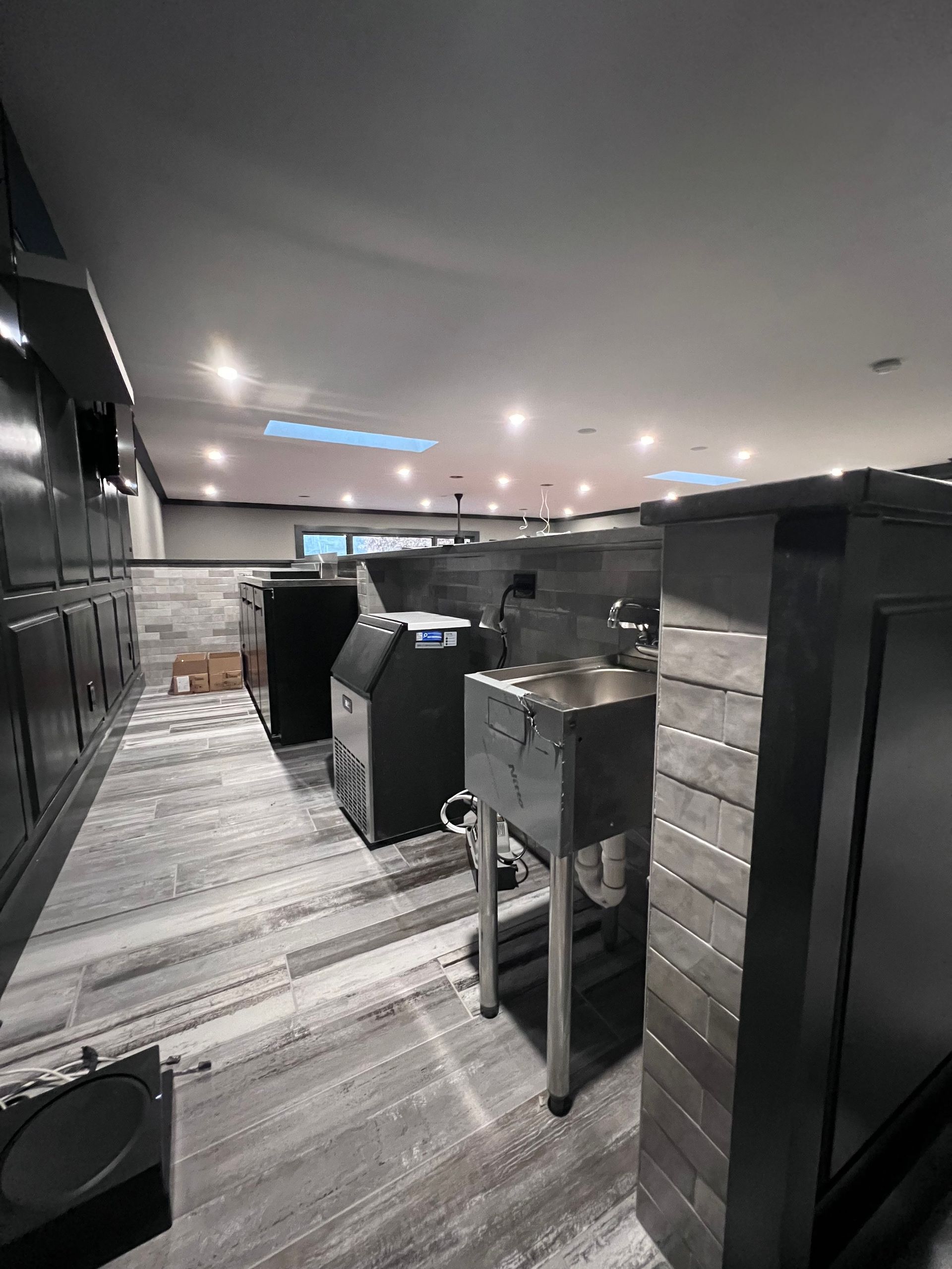 Commercial kitchen with stainless steel appliances, grey brick, and tiled floor under bright lights.