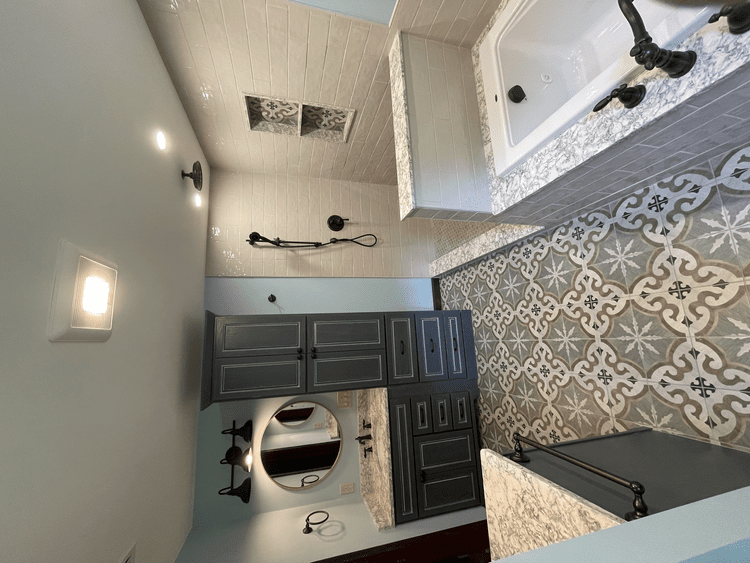 Bathroom with gray and white tile, dark cabinets, a round mirror, and a bathtub.