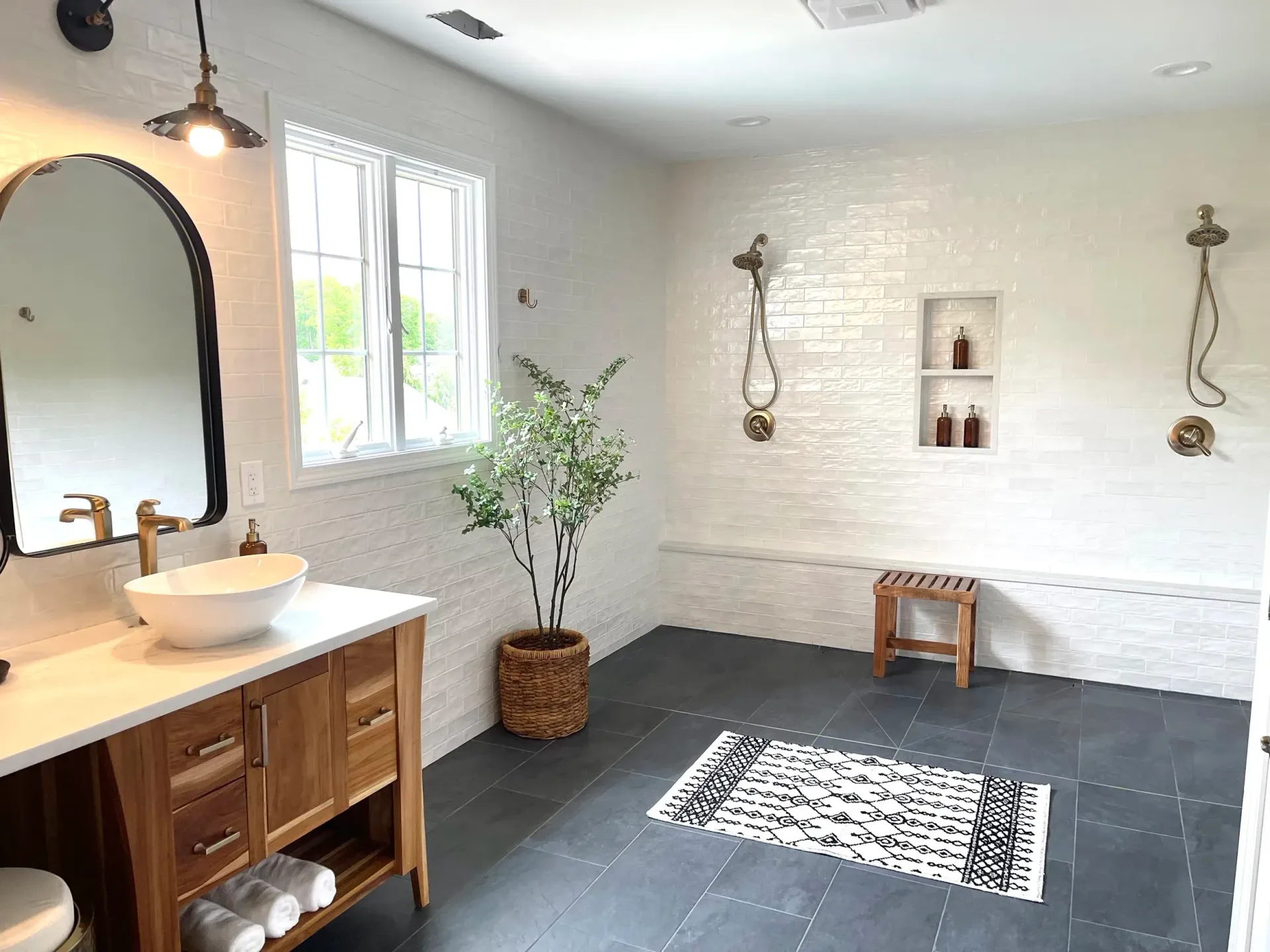 Bathroom with wood vanity, white brick walls, dark floor, shower with bench, arch mirror, window with curtain.