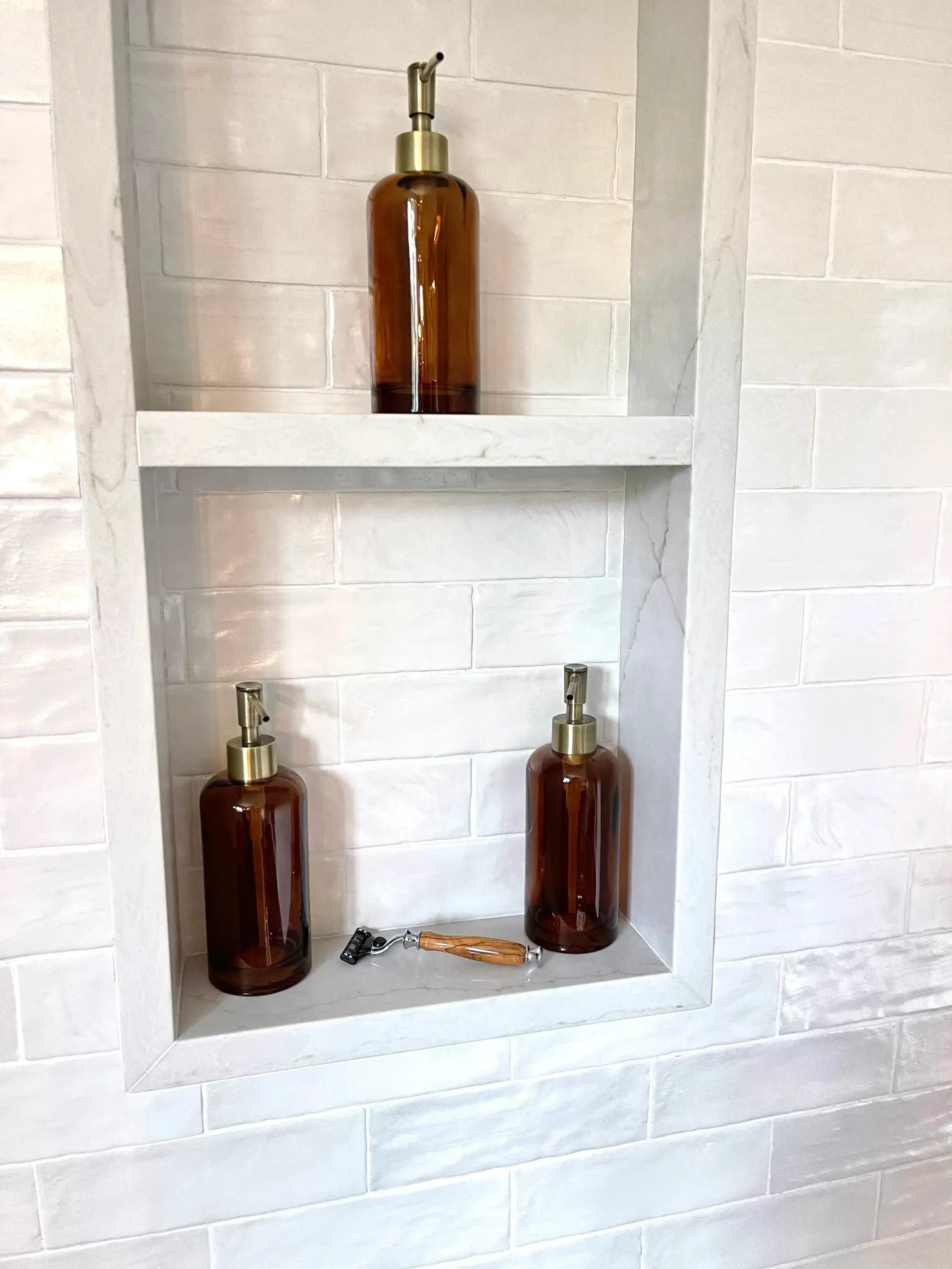 Three brown glass bottles with gold pumps in a recessed white tiled wall shelf. A razor lies below.